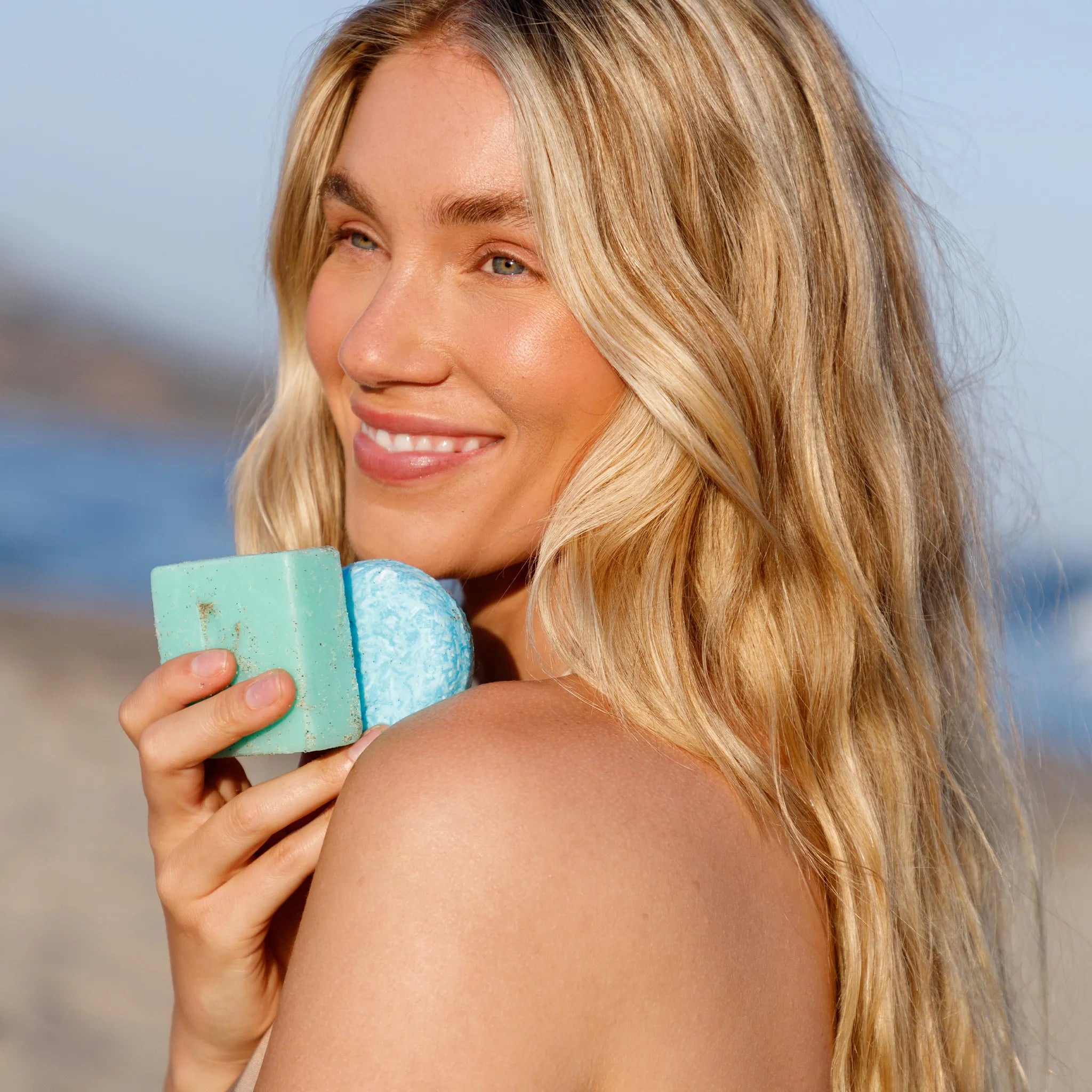Woman holding seaweed shampoo and soap against a beach background