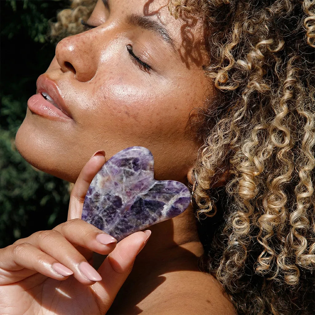 Woman holding a Seek Bamboo Amethyst Gua Sha stone against her face outdoors.