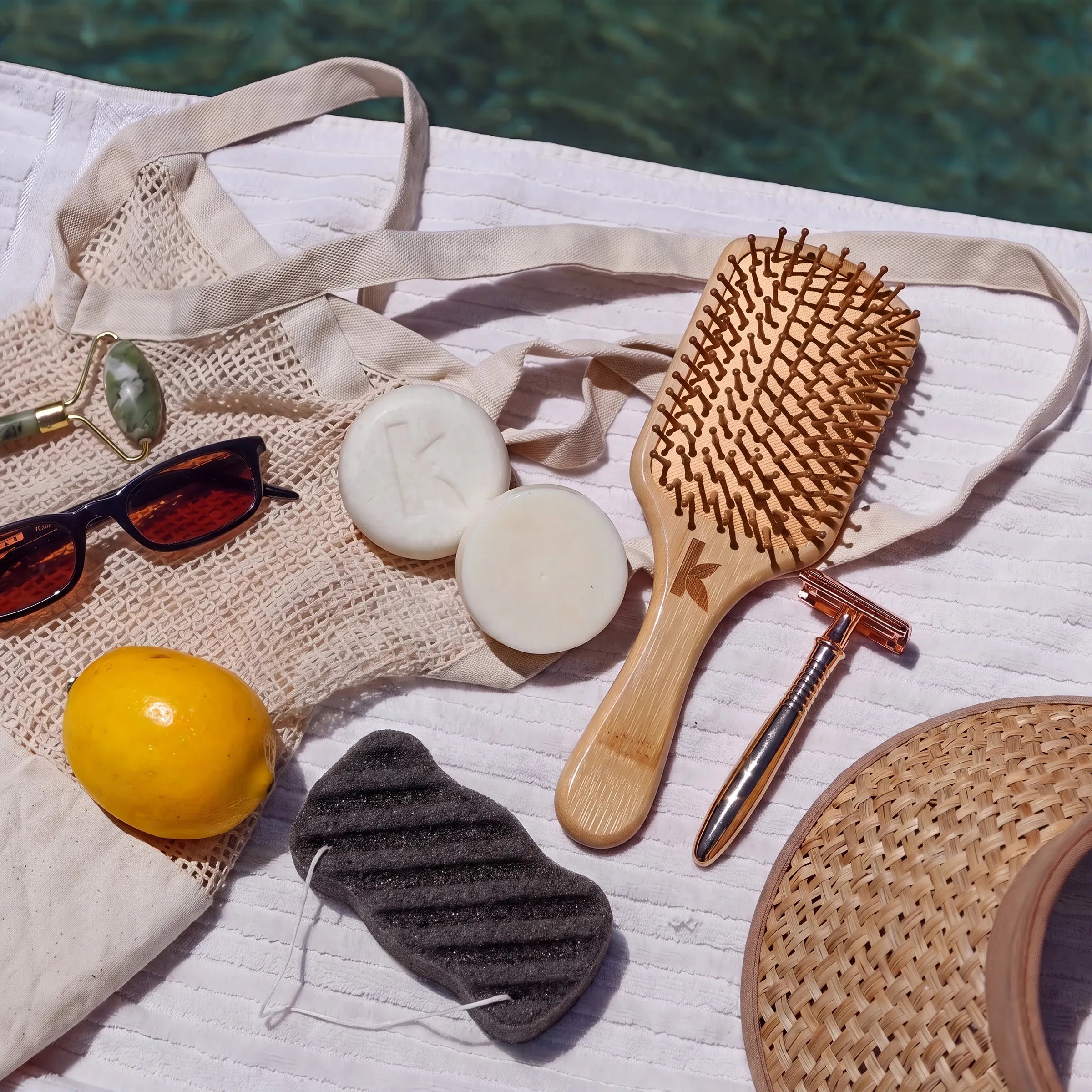 A styled flat-lay of beach and spa accessories on a white towel by the pool, including a woven tote bag, wooden hairbrush, rose gold razor, cream soap bars, sunglasses, a yellow lemon, a charcoal konjac sponge, and a straw hat.