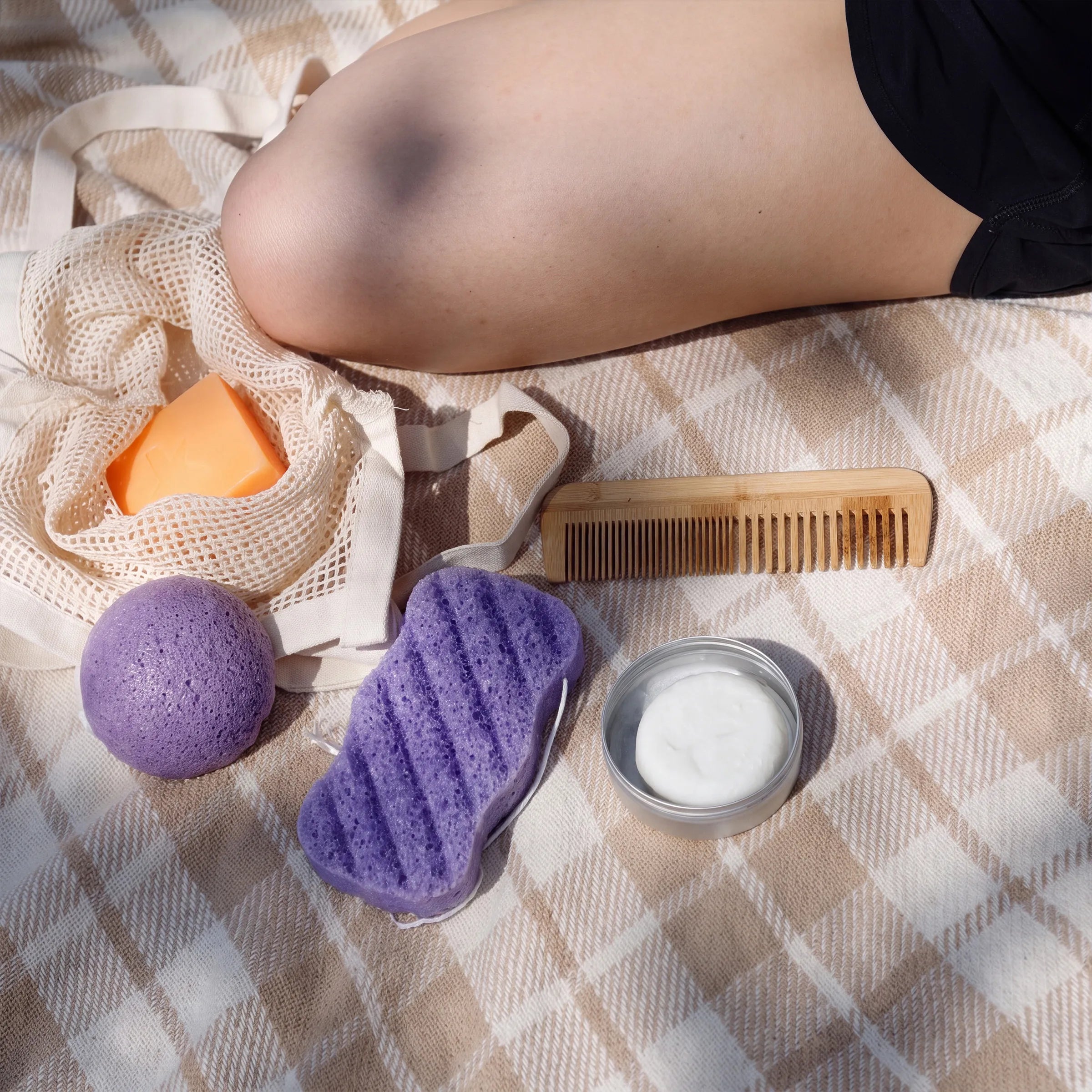 A styled flat-lay of a skincare set on a beige and white plaid blanket, featuring a lavender textured sponge, a purple bath bomb, an orange soap bar, a bamboo comb, and a small jar of white cream next to a reusable mesh bag.