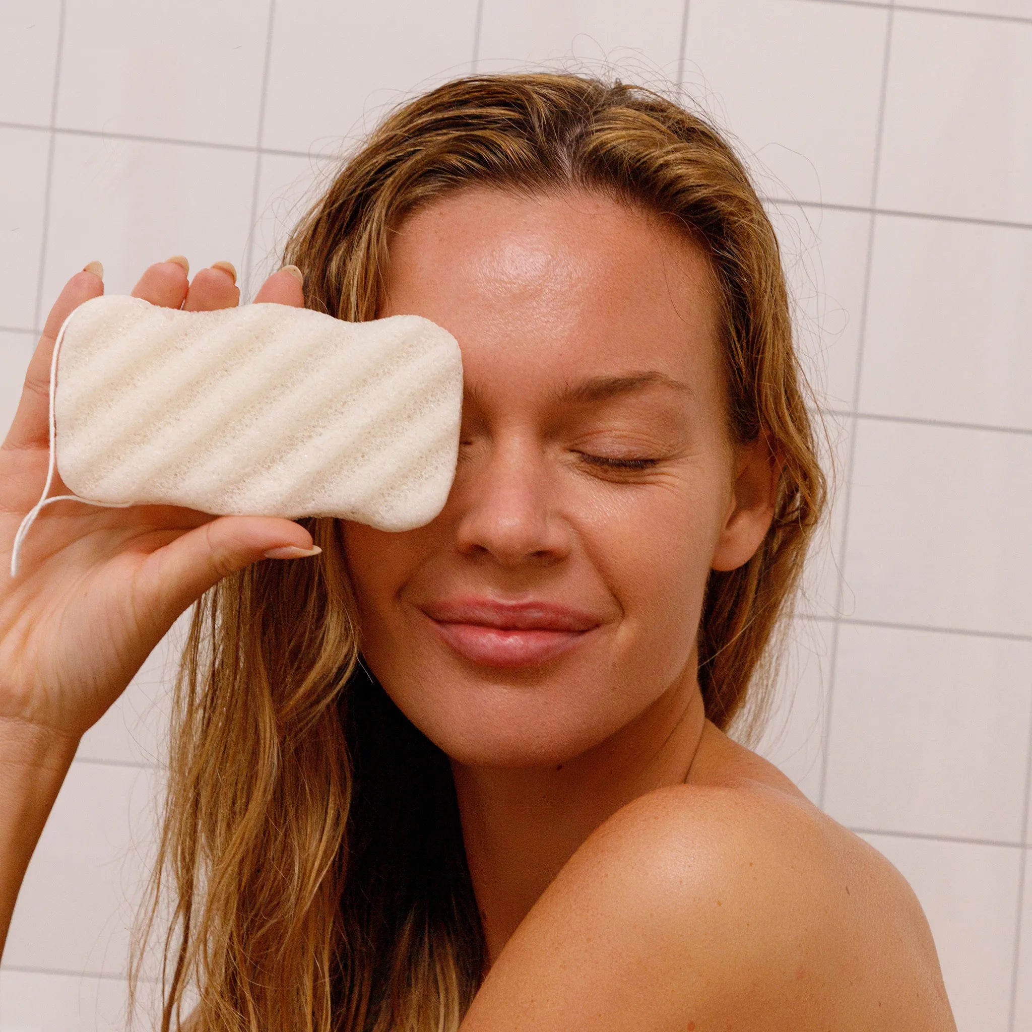 Woman holding unscented konjac sponge to her face against a tiled bathroom background.