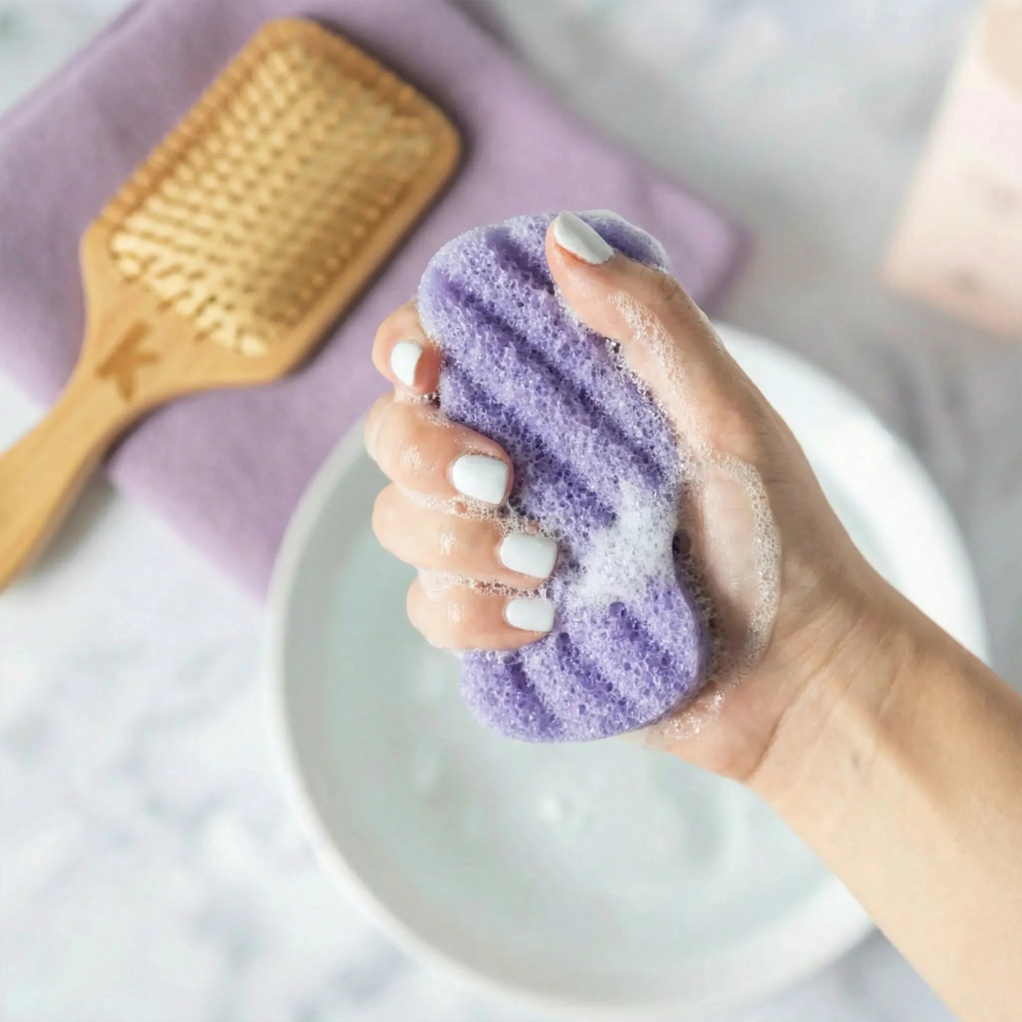 Hand holding a lavender konjac body sponge with a brush and towel in the background