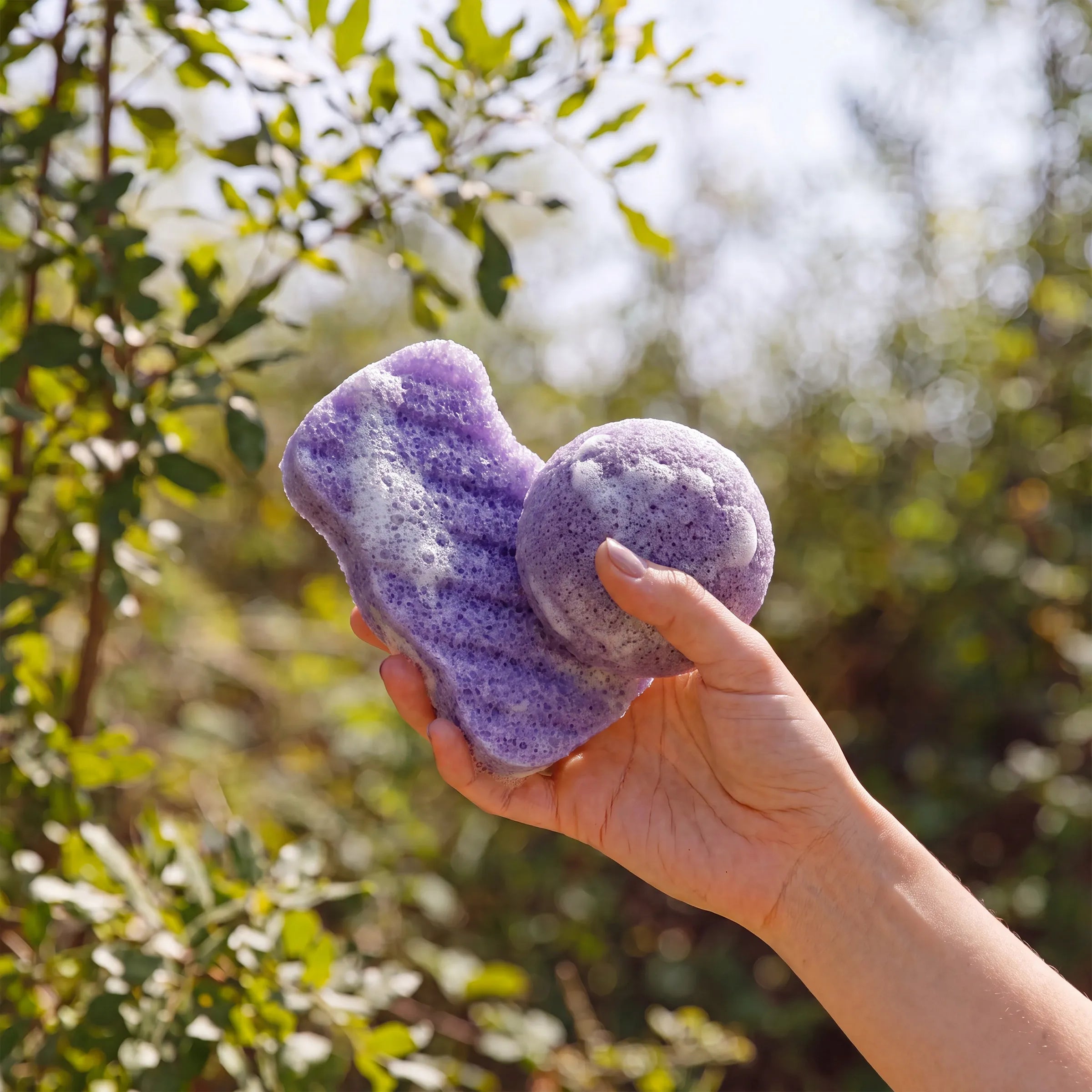 A hand holding lavender textured exfoliating konjac scrub sponge outdoors with green foliage in the background.