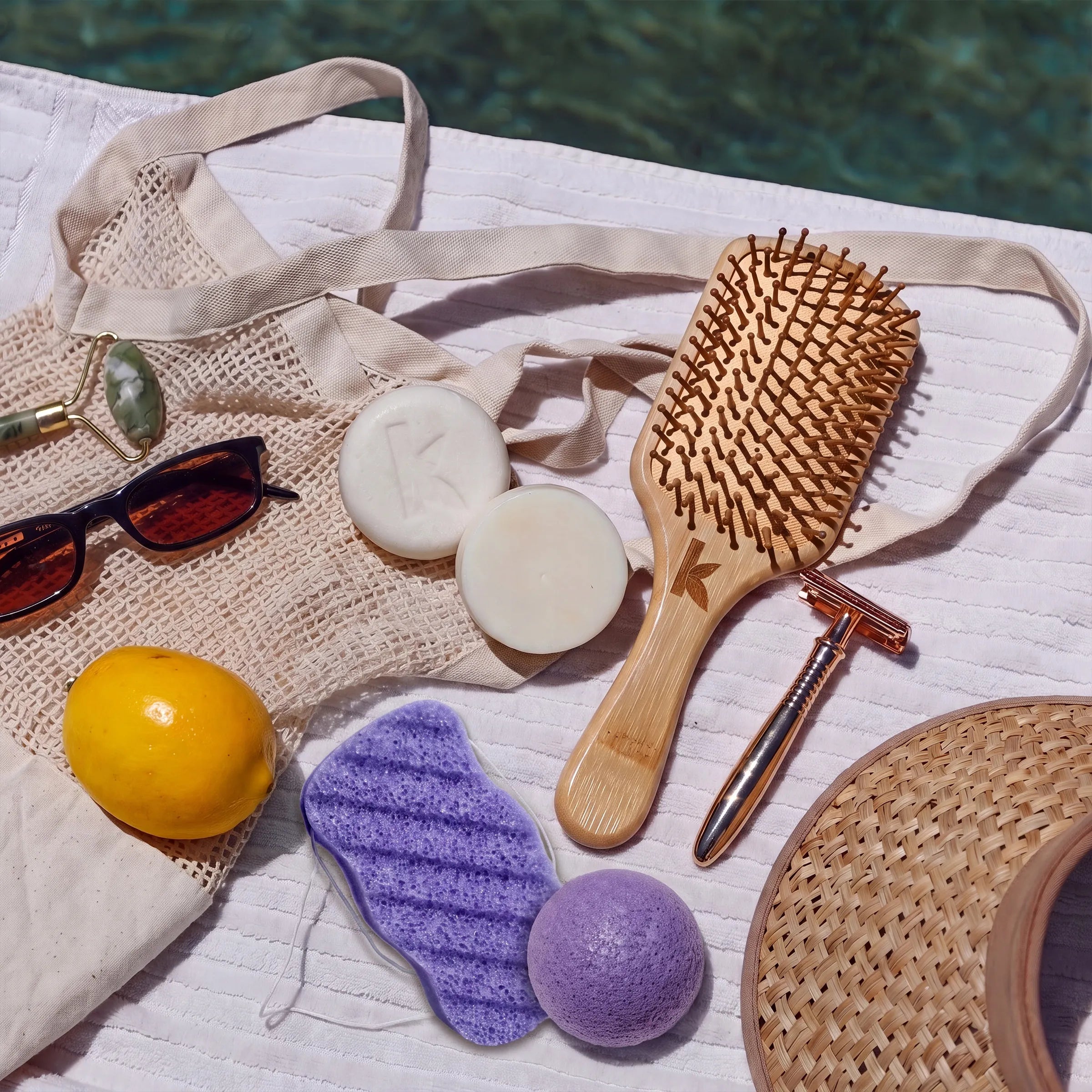 A styled flat-lay of wellness and beauty accessories on a white textured surface, including a wooden hairbrush, a metal safety razor, two round white bars of soap, a lavender konjac sponge, a yellow lemon, black sunglasses, and a woven straw hat.