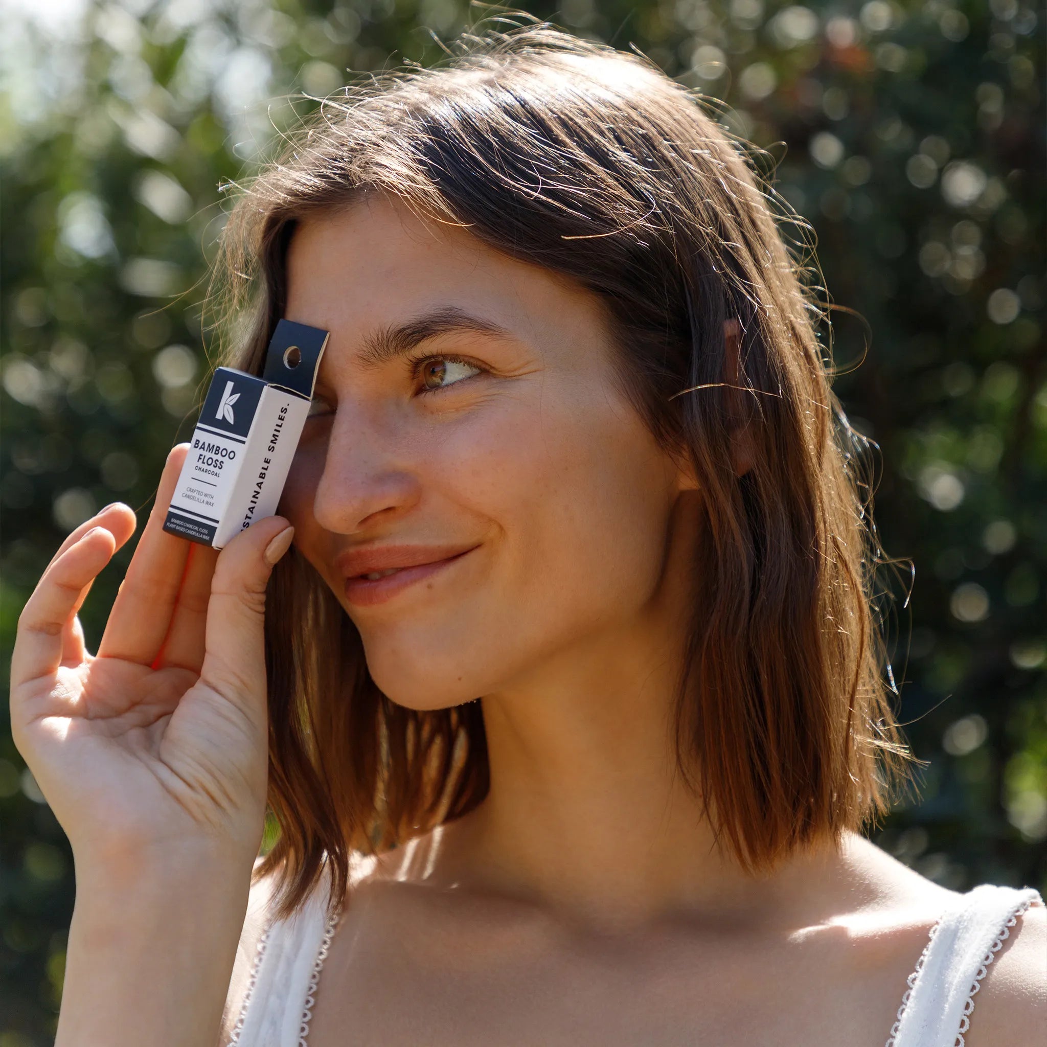 A woman outdoors holds a bamboo floss box near her eye. The box has a white label with a black logo and the text 'Seek Bamboo'.