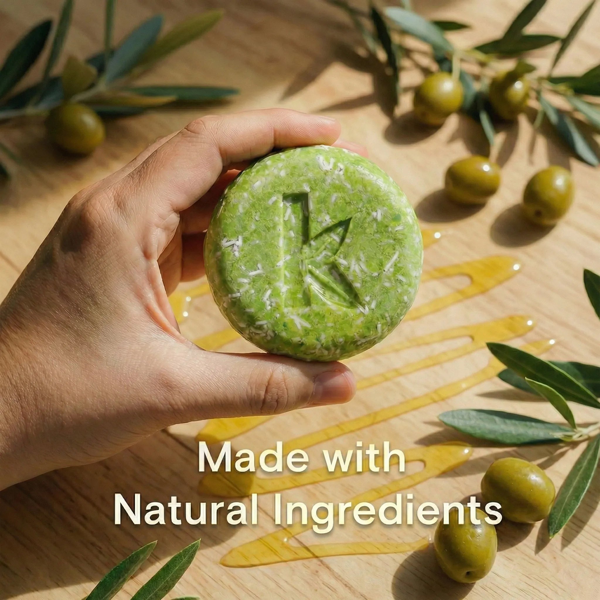 Hand holding a olive oil shampoo bar with olive leaves and oil on a wooden background, labeled 'Made with Natural Ingredients'.