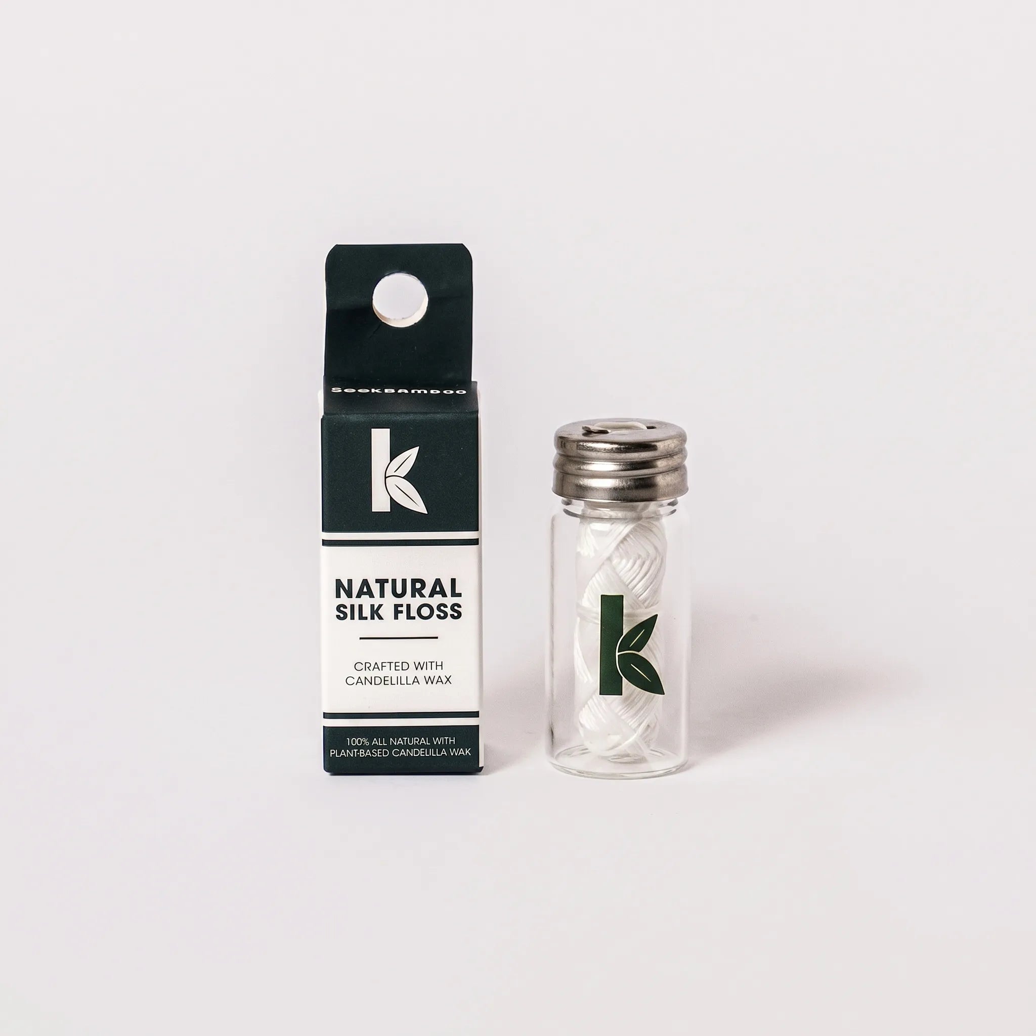 A small glass jar of silk floss with a silver metal lid stands next to its black and white retail box on a clean white background. The packaging features a green leaf logo and the text 'NATURAL SILK FLOSS' and 'CANDELILLA WAX'.