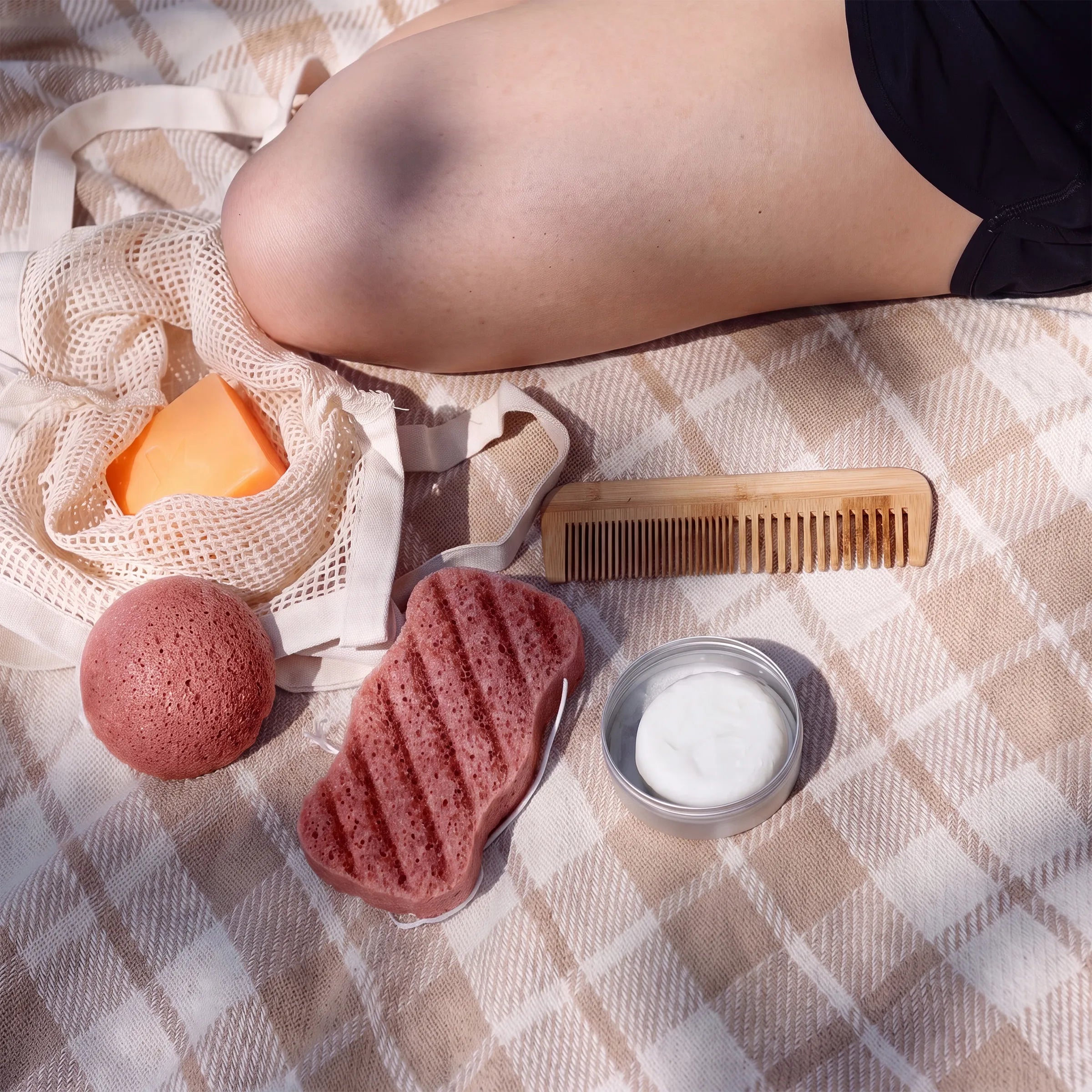 A styled flat-lay of skincare and bath accessories arranged on a beige plaid blanket, with a person's knee visible in the upper right. The set includes a mesh soap bag with orange soap, a red clay sponge, a striped pink scrub block, a small jar of white cream, and a wooden comb.