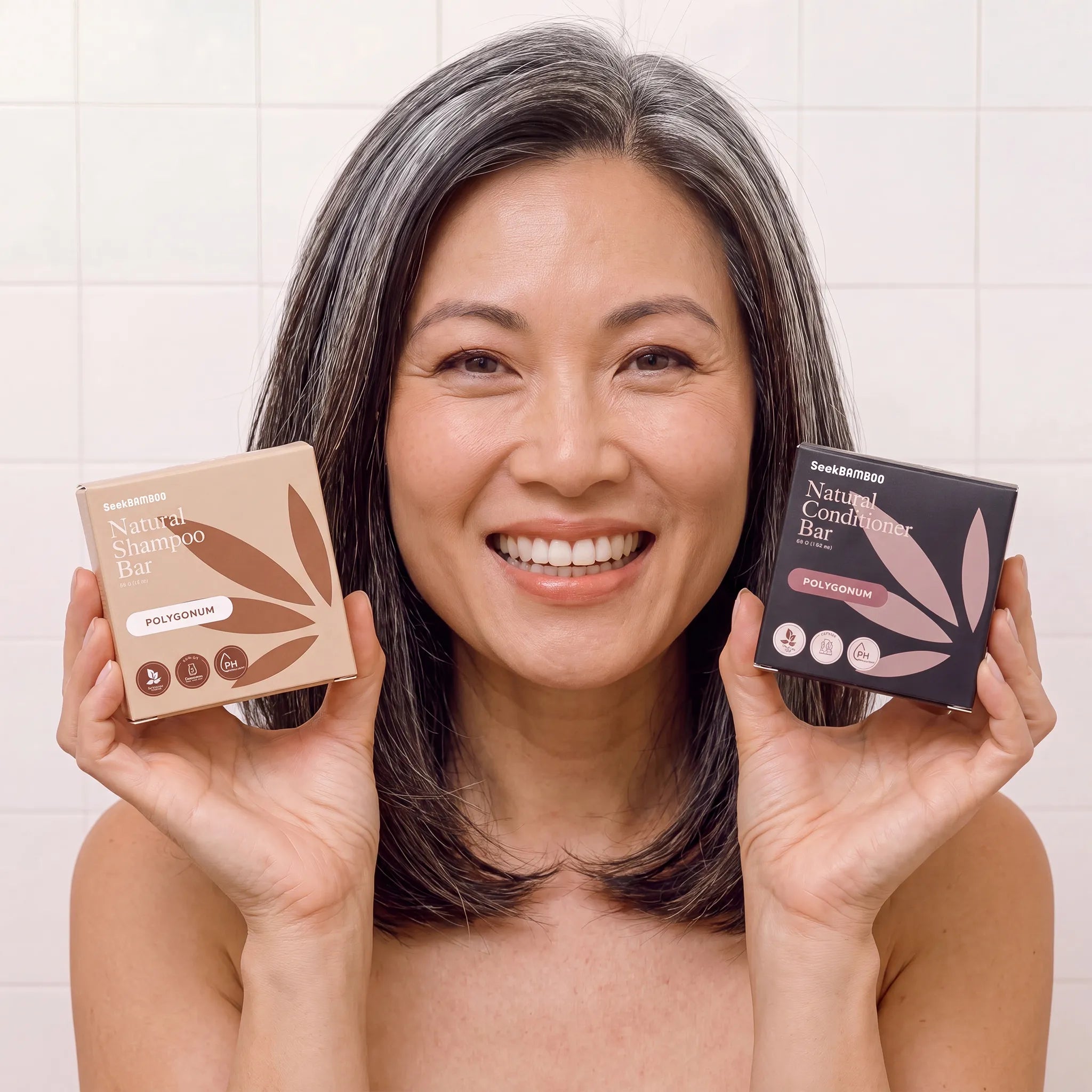 A smiling woman with gray hair holds up two polygonum seek bamboo hair care bars in front of her chest.