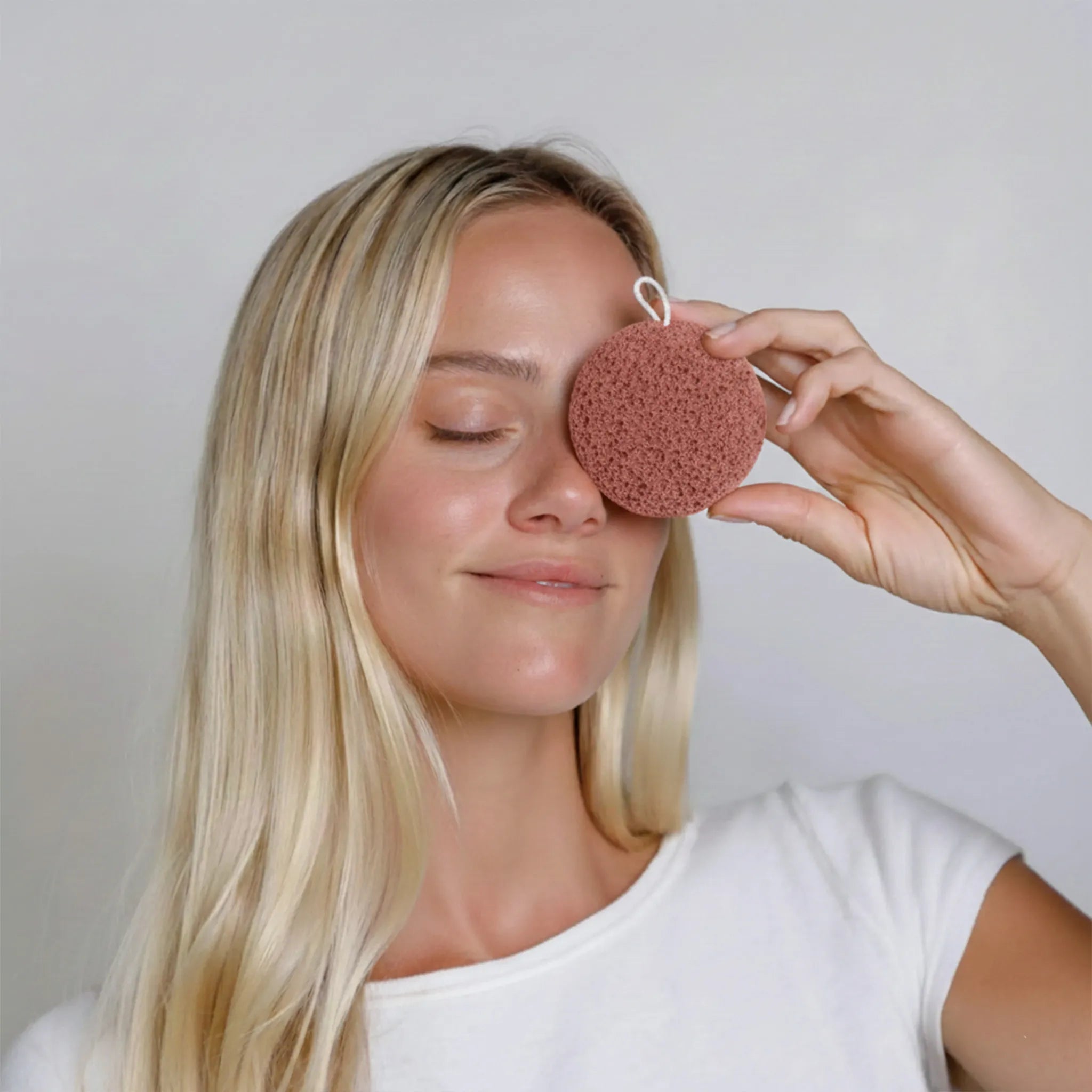 Woman using a red clay konjac sponge on her face against a plain background