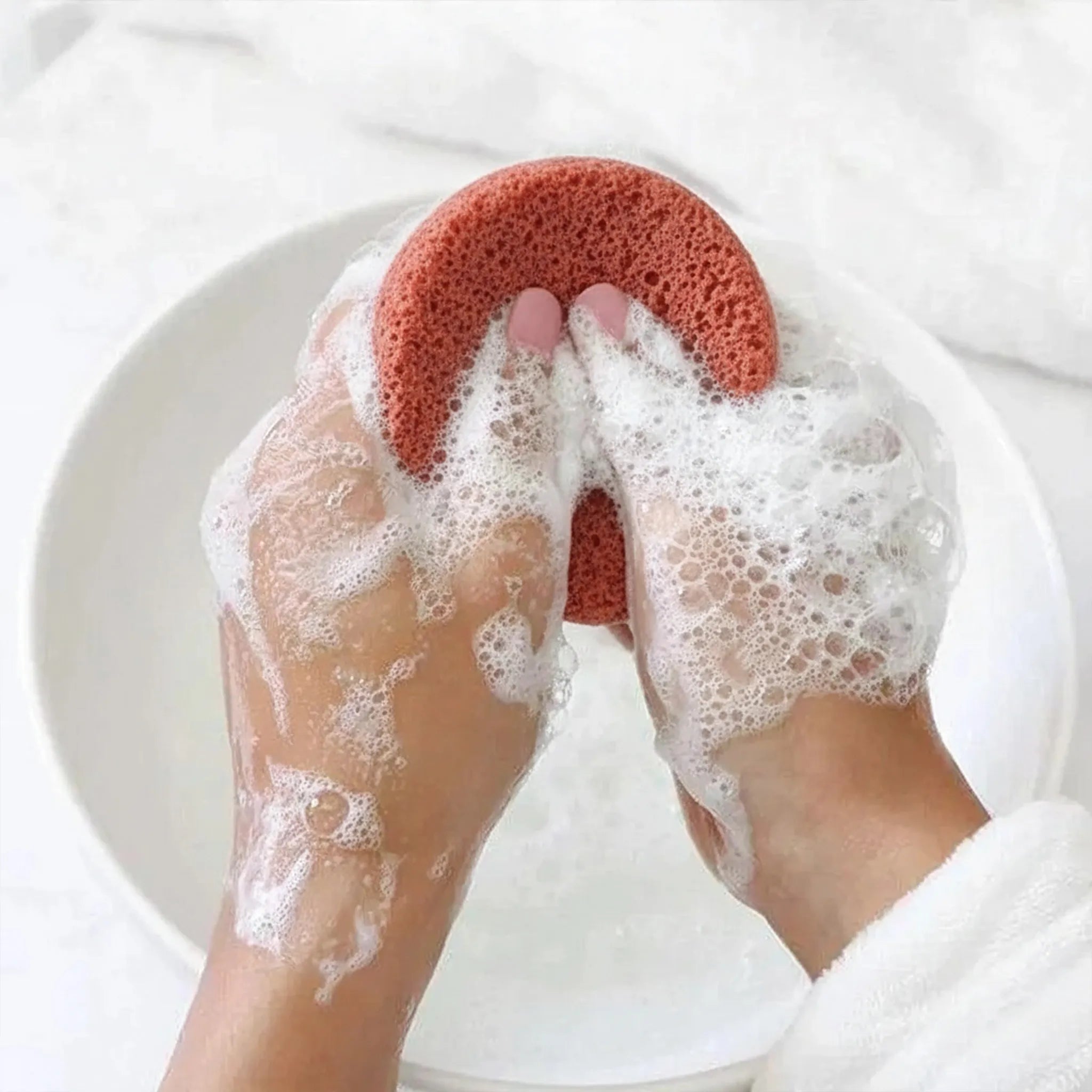 Person using a red clay konjac sponge to scrub their hands with soap suds on a white background