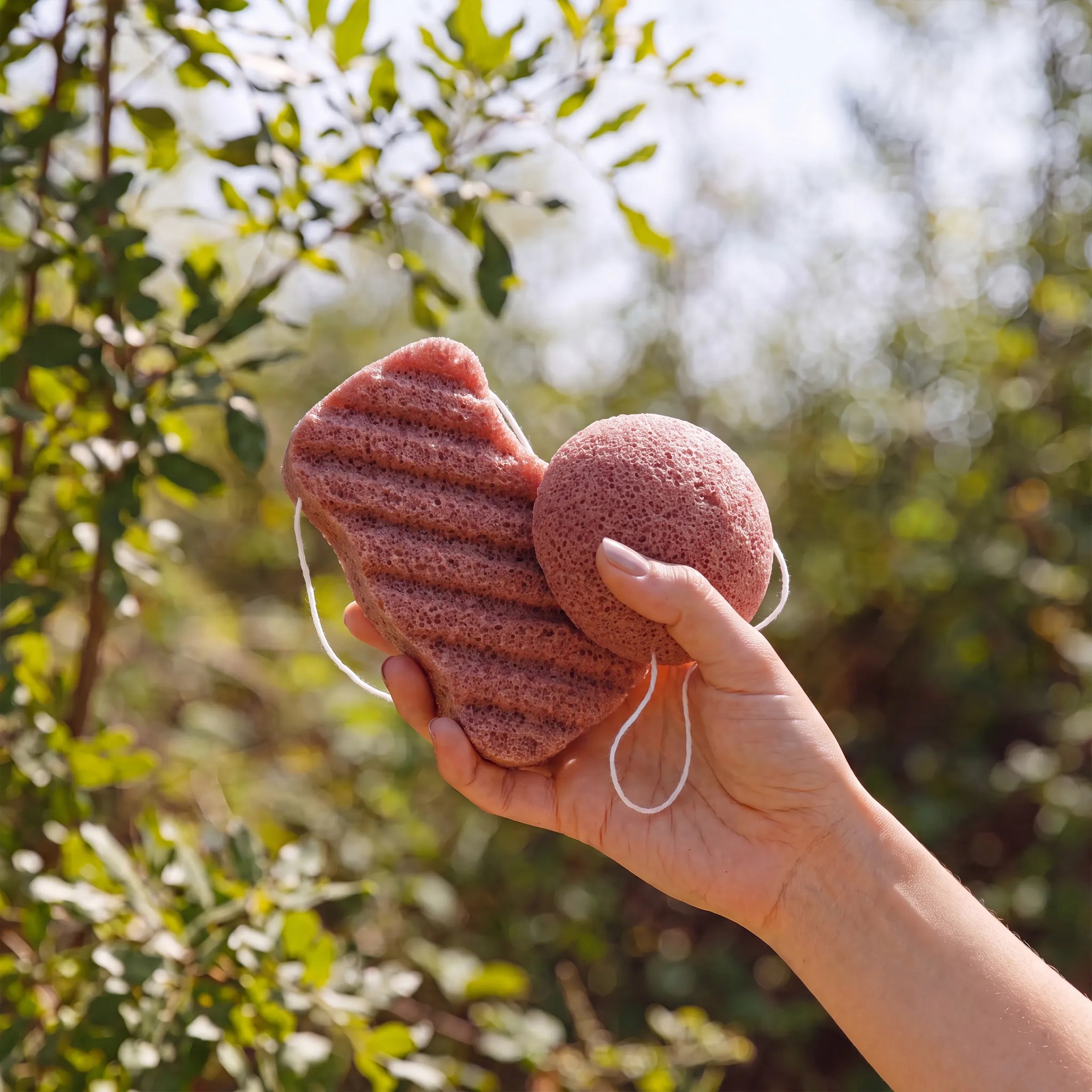 A hand holding two red clay textured bath sponge shapes outdoors against a blurred green foliage background.