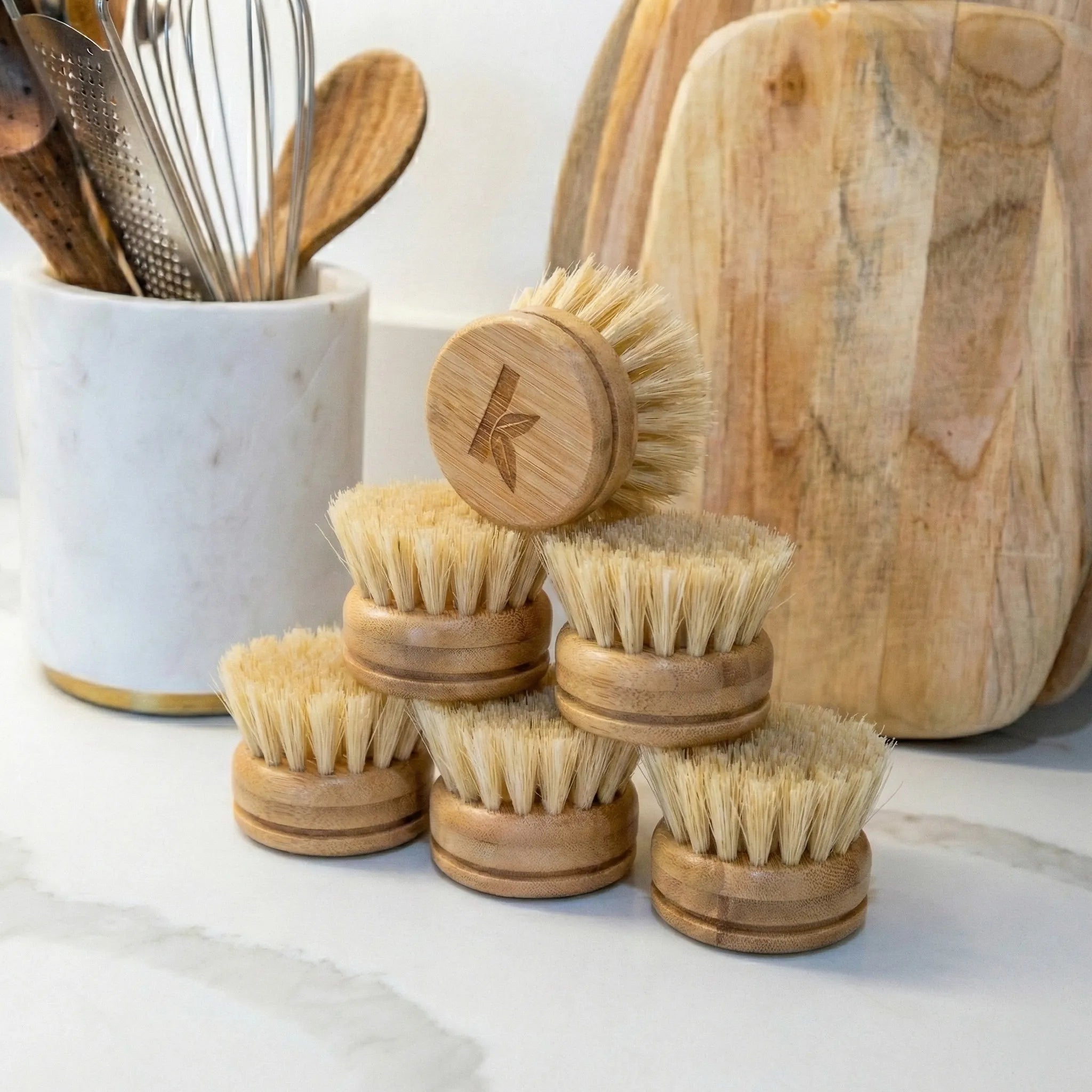 Set of bamboo dish brushes on a marble surface with kitchen utensils in the background.