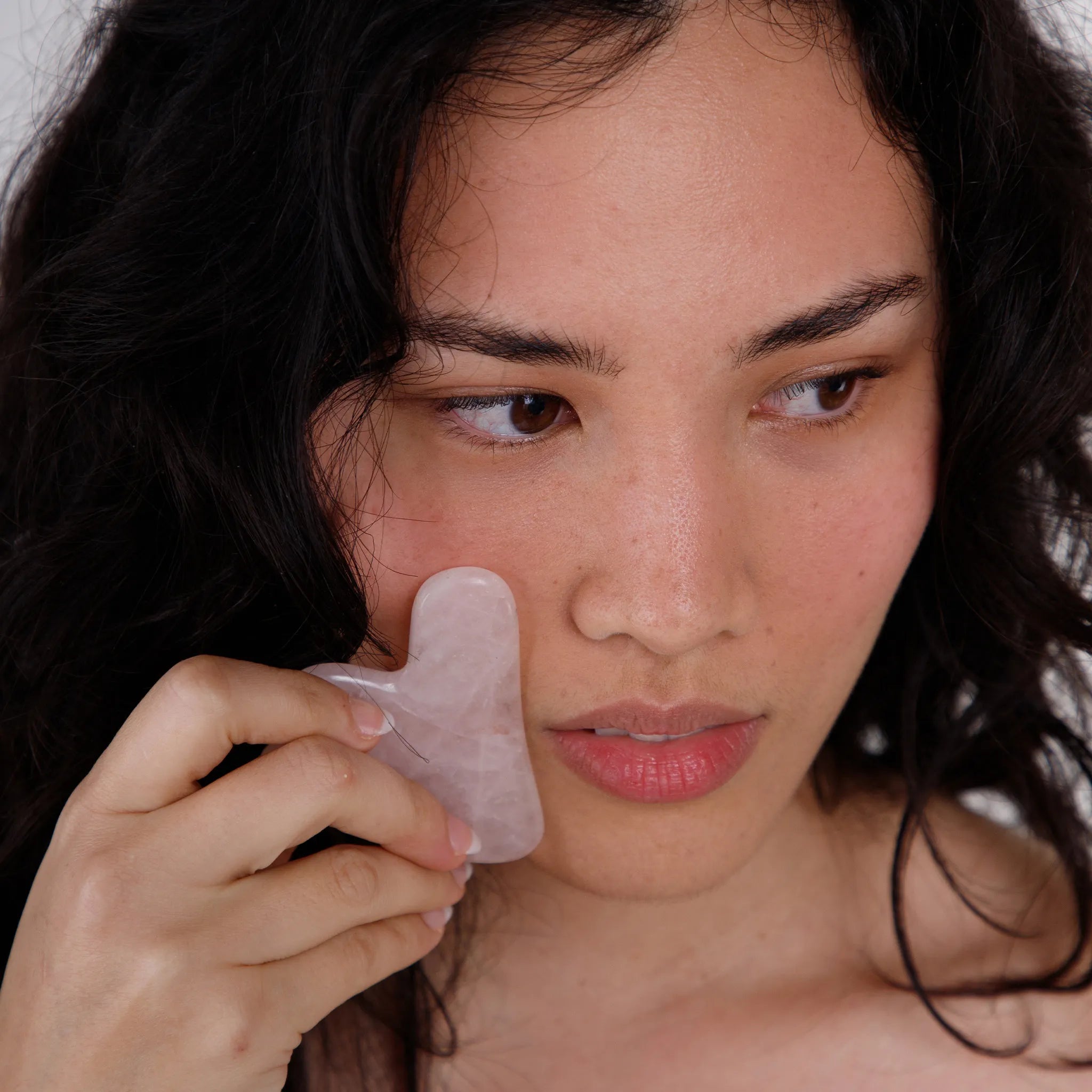 Woman using a pink rose quartz gua sha on her face against a neutral background