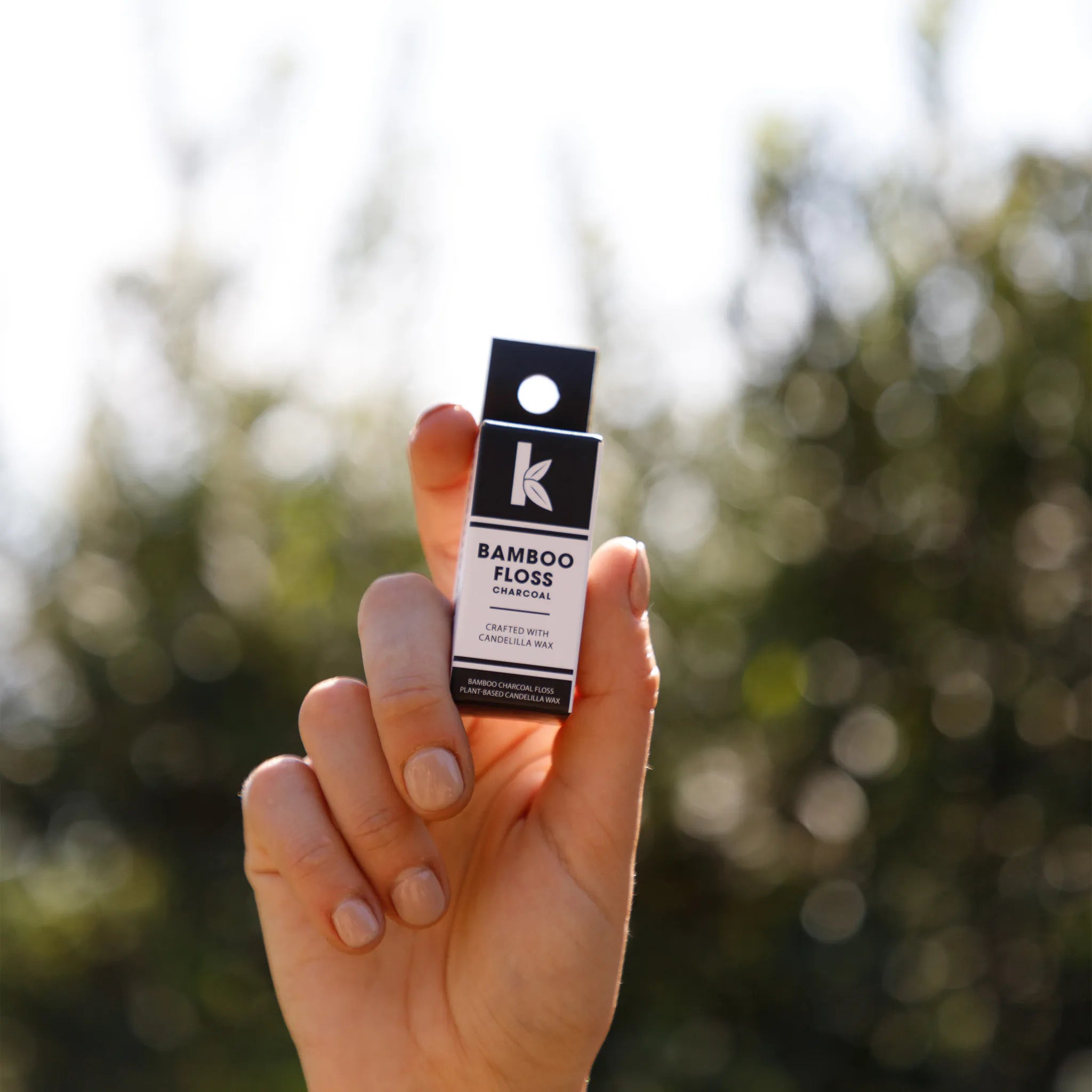 A hand holding a small black and white box of Seek Bamboo Floss against a blurred outdoor background of green foliage and bright sunlight.