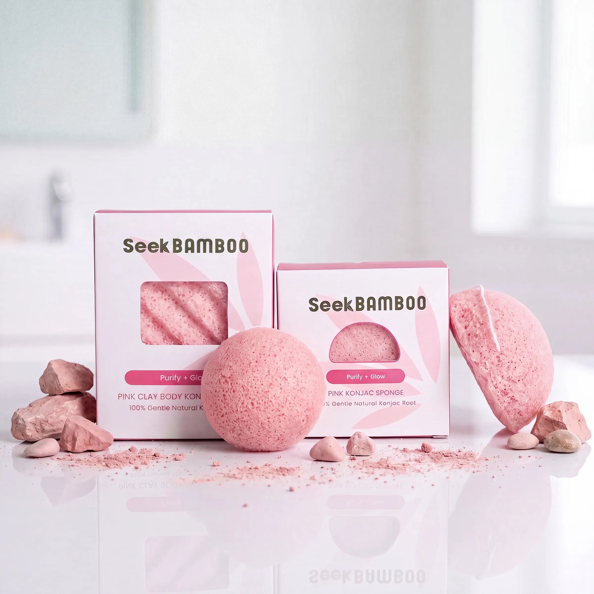 Two pink konjac spongesfrom Seek bamboo, including a pink clay body foam box and a pink konjac sponge box, displayed with their corresponding pink bath bombs and small pink stones on a reflective white surface.