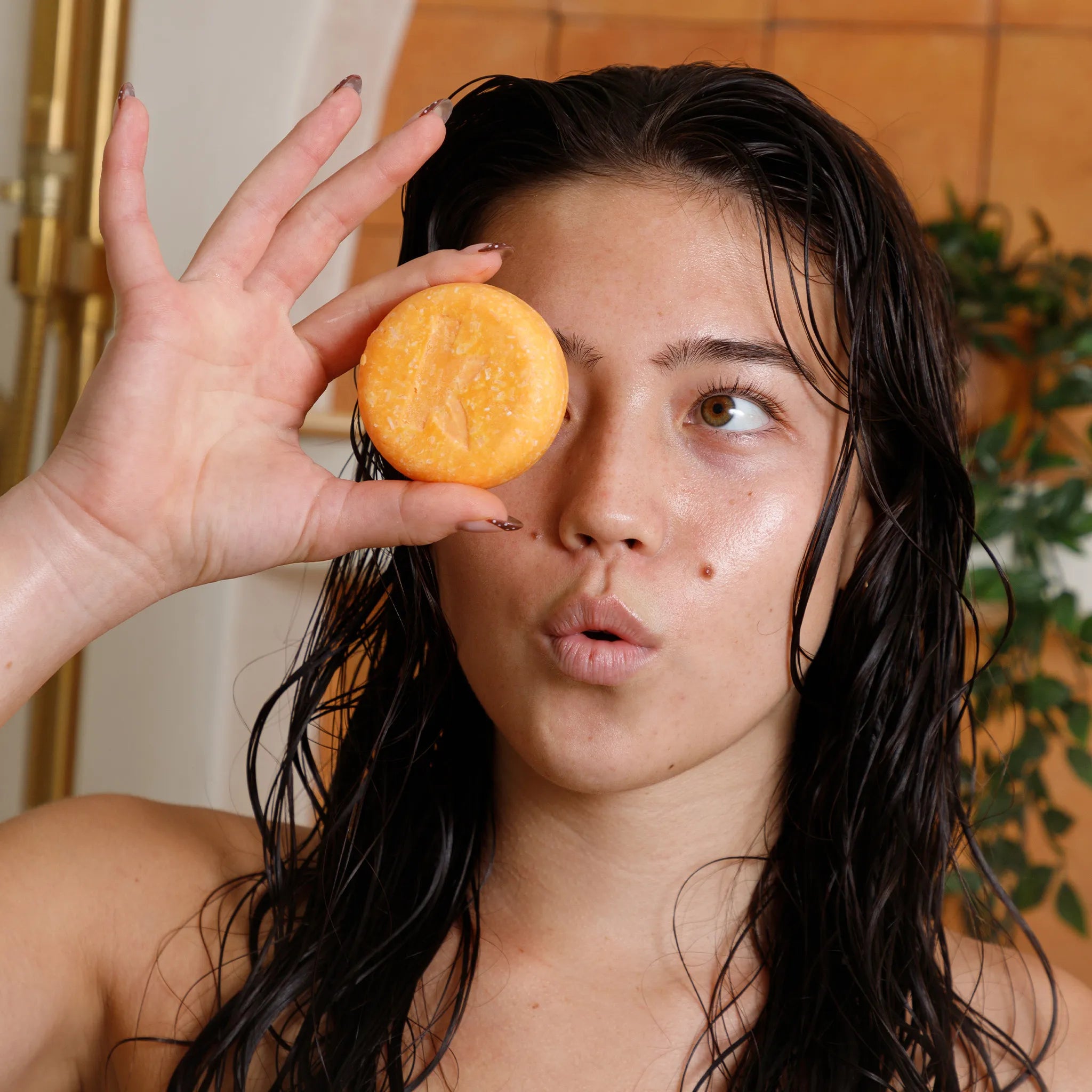 Woman with wet hair holding a honeyt shampoo bar near her face in a bathroom setting.