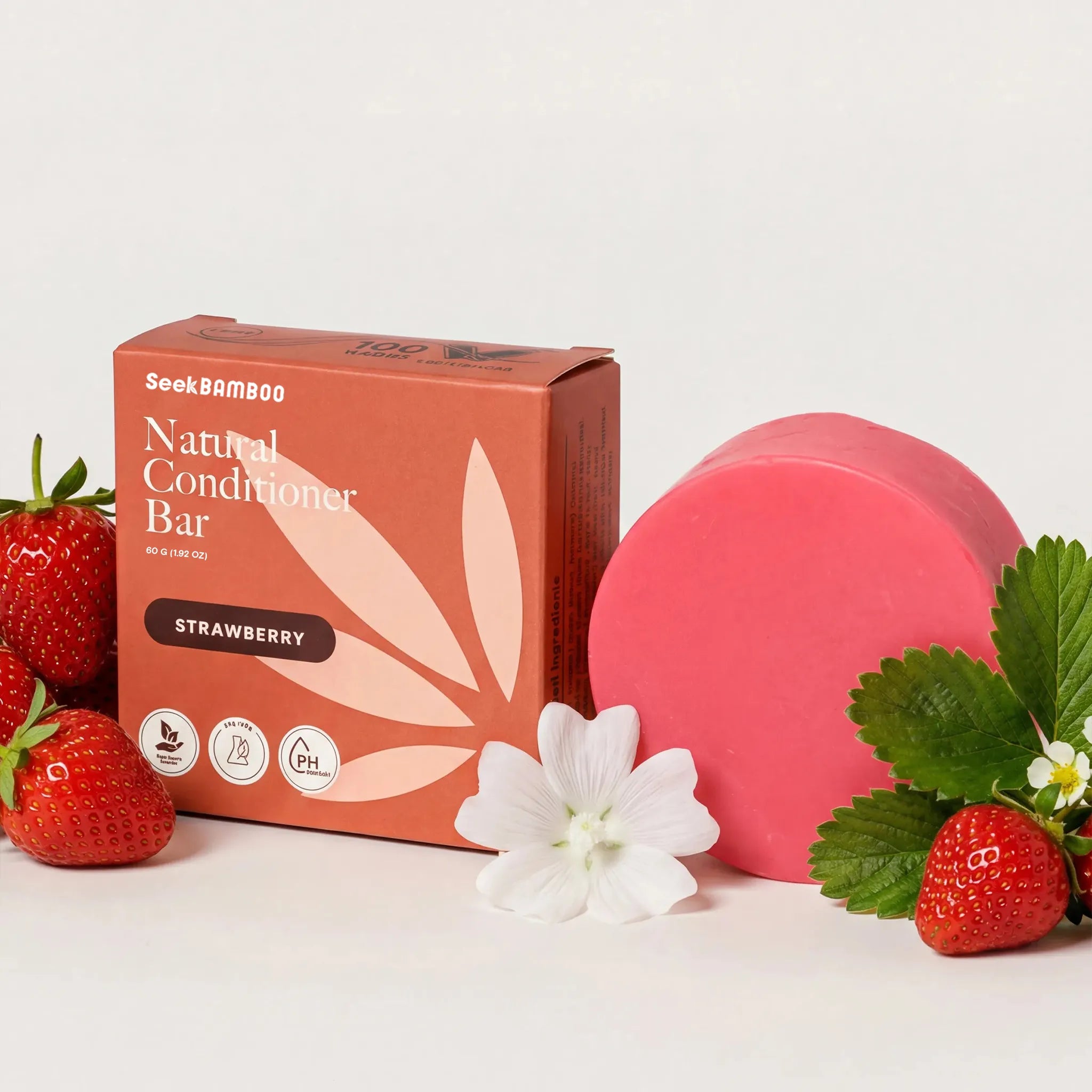 a strawberry conditioner bar, shown with its terracotta-colored packaging box, a pink conditioner bar, fresh strawberries, green leaves, and a white flower on a clean white background.