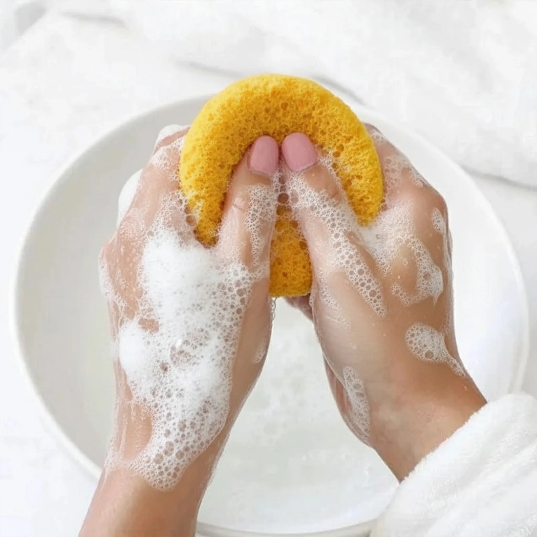 Hands scrubbing a turmeric konjac sponge with soap suds in a white bowl.