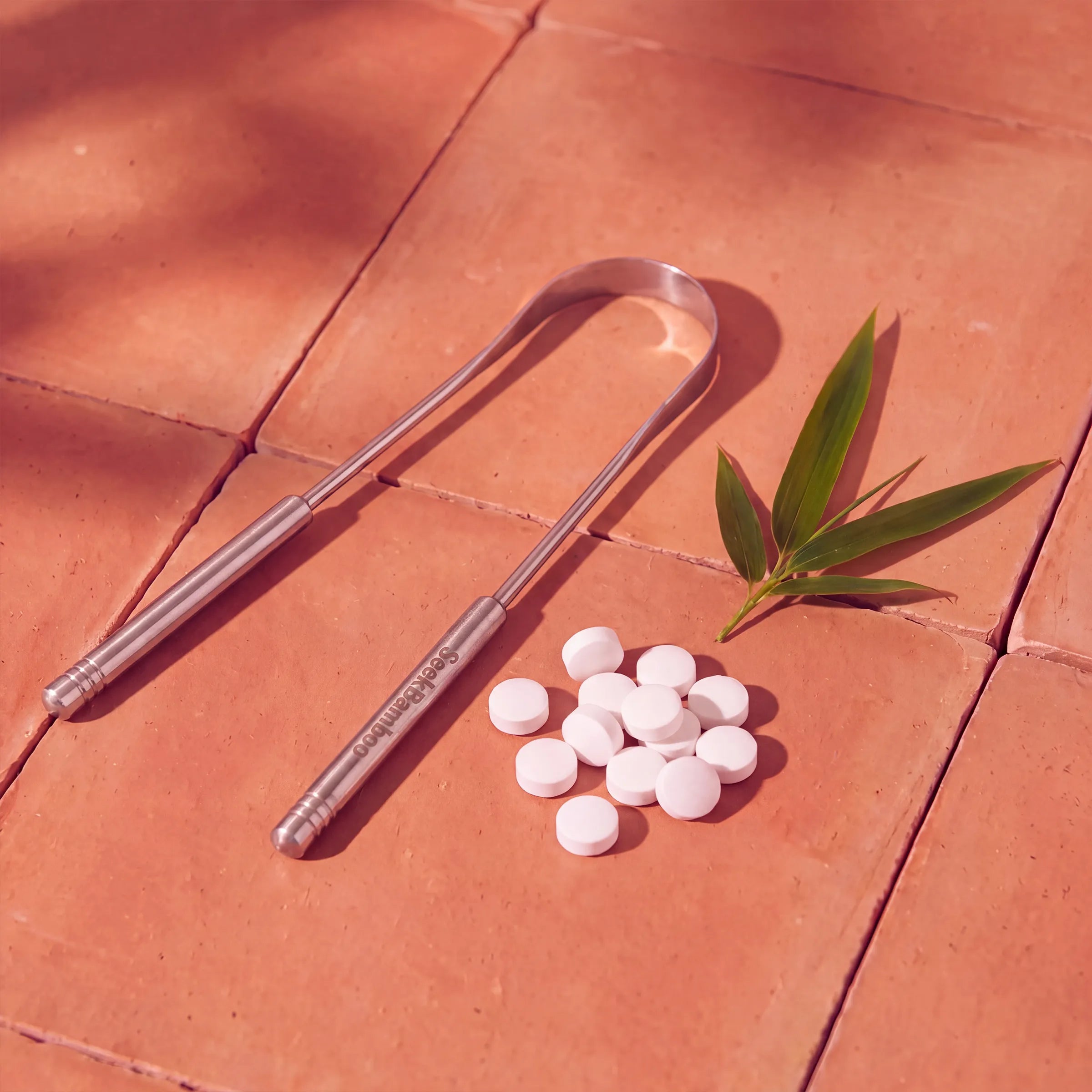 A stainless steel tongue scraper with a bamboo handle lies on terracotta tiles next to a small pile of white tablets and a green bamboo leaf.