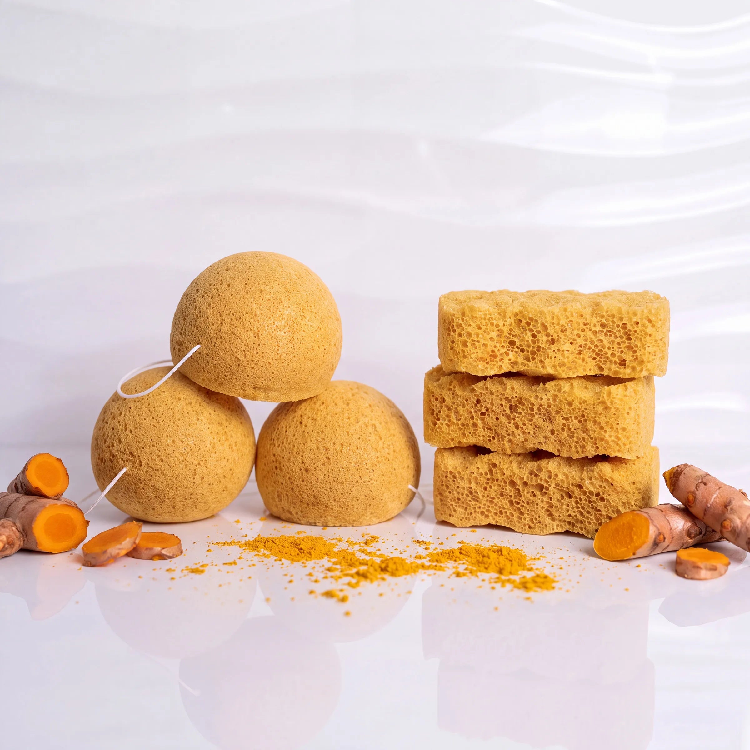Three round and three rectangular bath sponge blocks in a warm golden-brown color, arranged with sliced turmeric roots and loose turmeric powder on a glossy white surface against a softly textured white background.