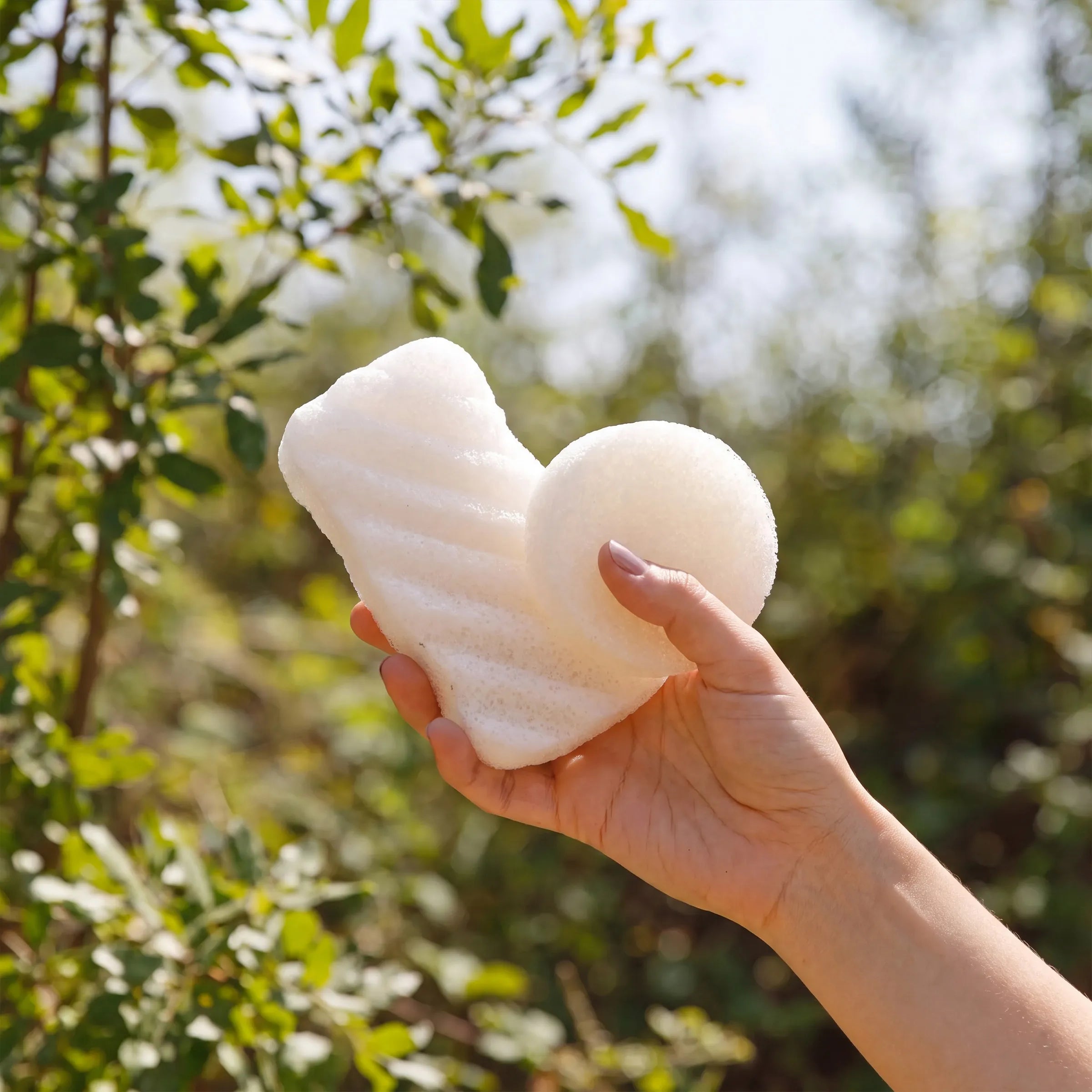 A hand holding a white, unscented konjac cleaning sponge with a round scrubber attachment, set against a blurred outdoor background of green foliage and sunlight.