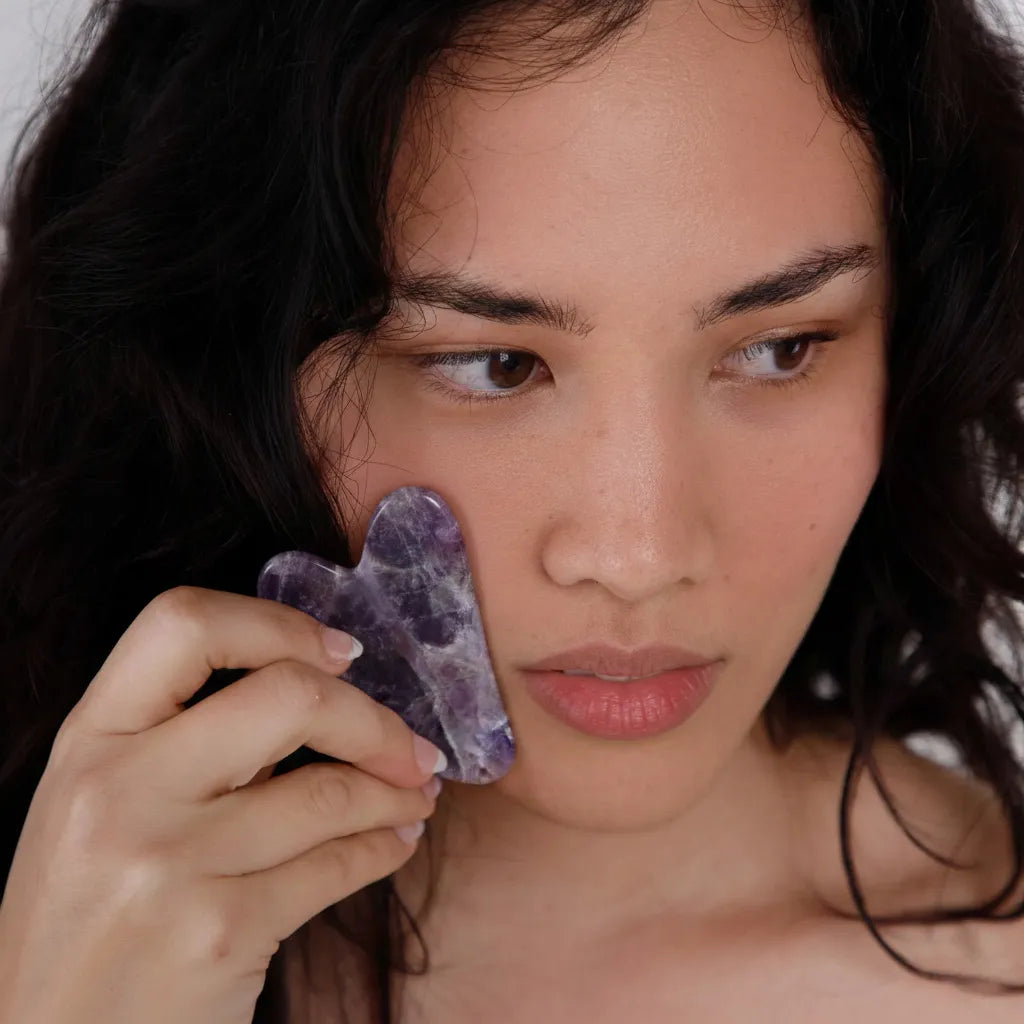 Woman holding a Amethyst Gua Sha tool near her face against a neutral background