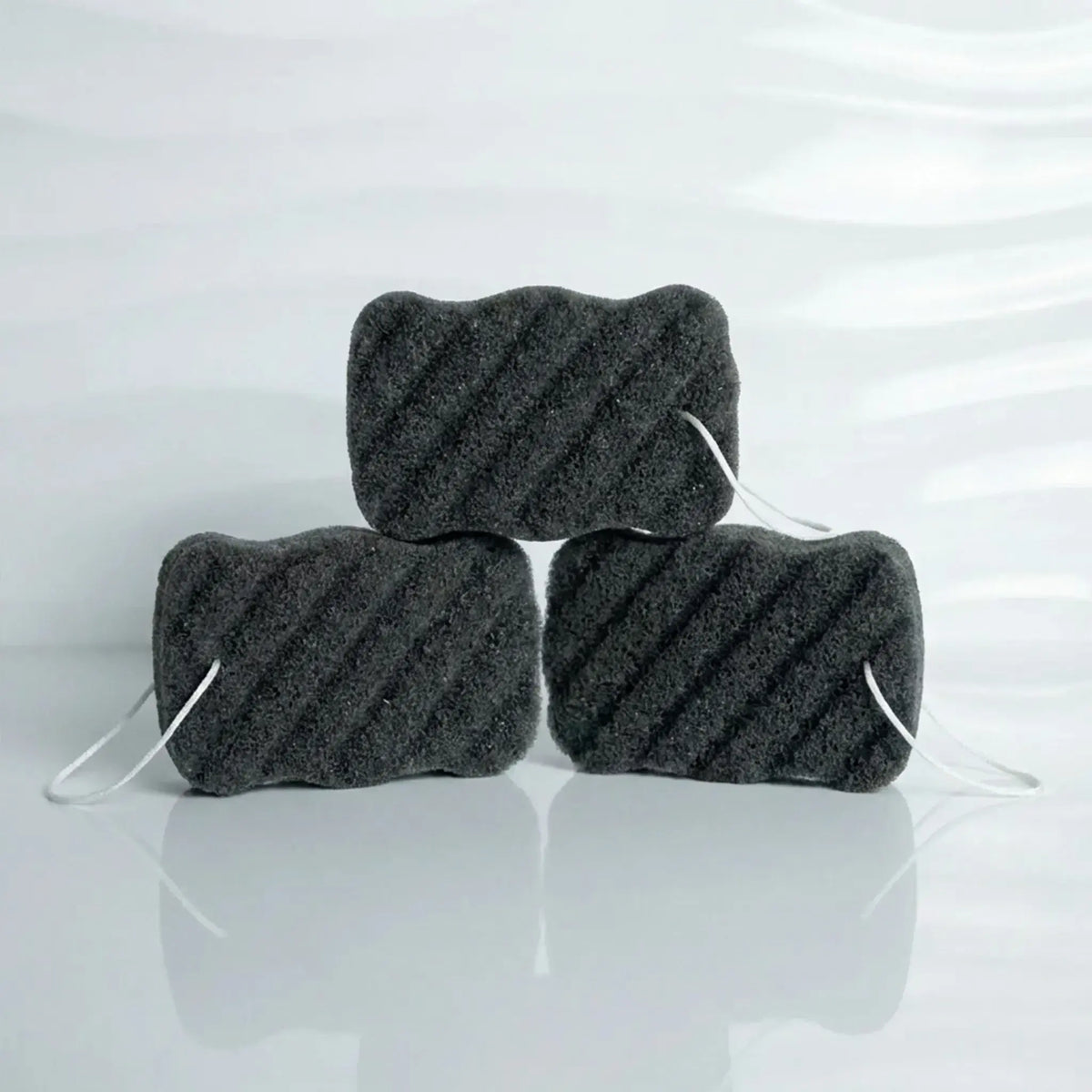 Three charcoal konjac body sponge blocks with white elastic ties, arranged in a pyramid stack on a reflective white surface.