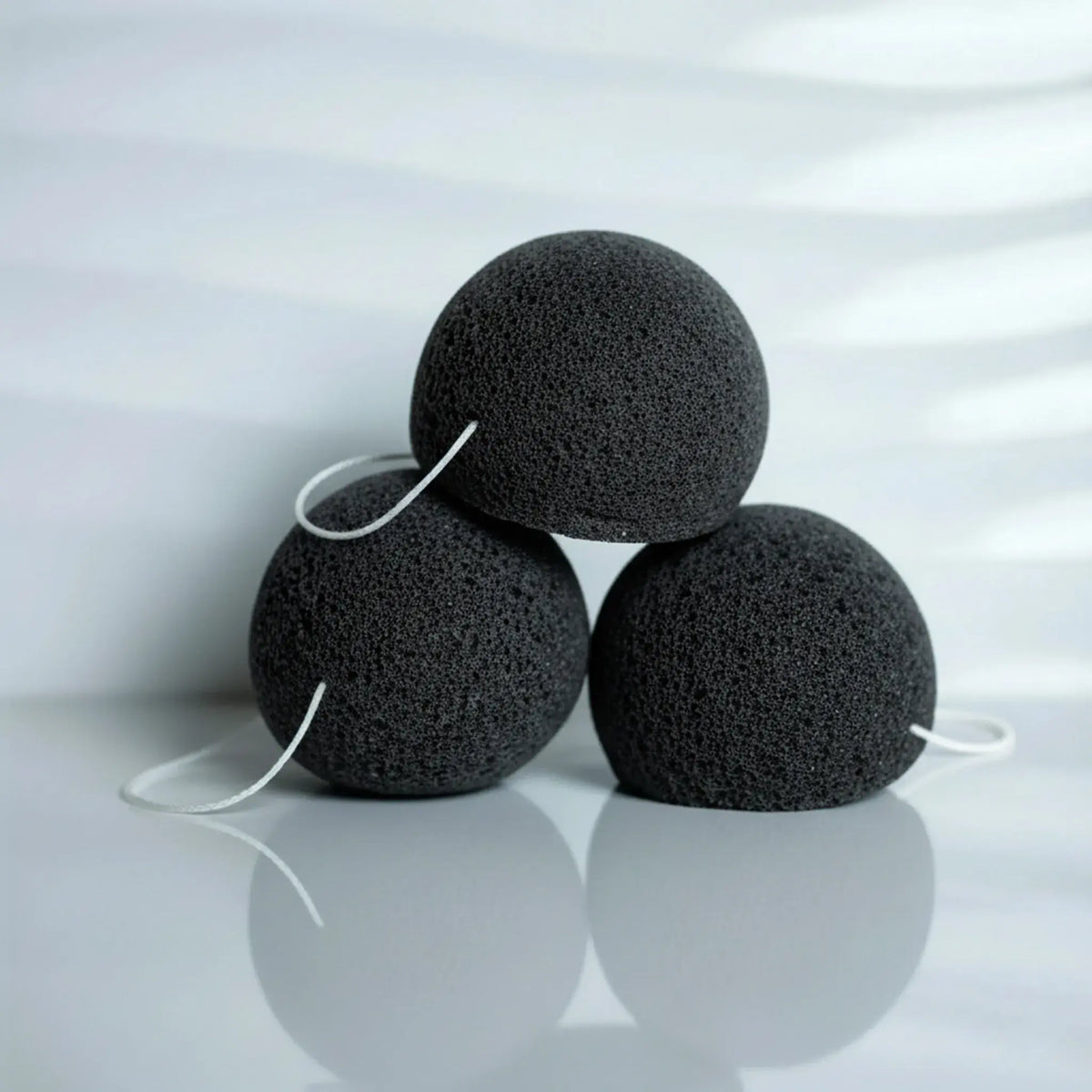 Three charcoal konjac sponges with white elastic loops, arranged in a pyramid on a reflective light gray surface.