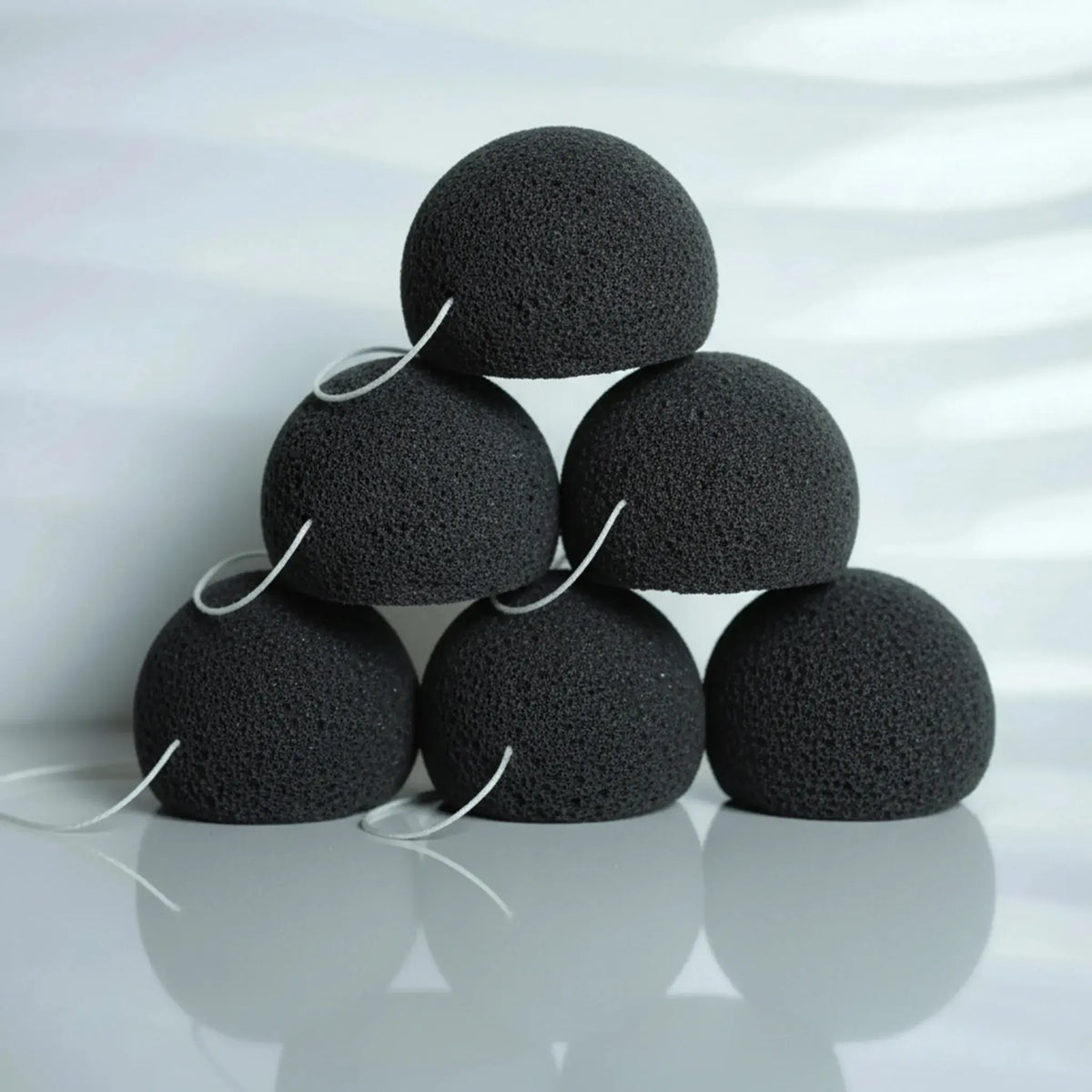 Six charcoal konjac sponges with white elastic ties, stacked in a pyramid arrangement on a reflective light gray surface.