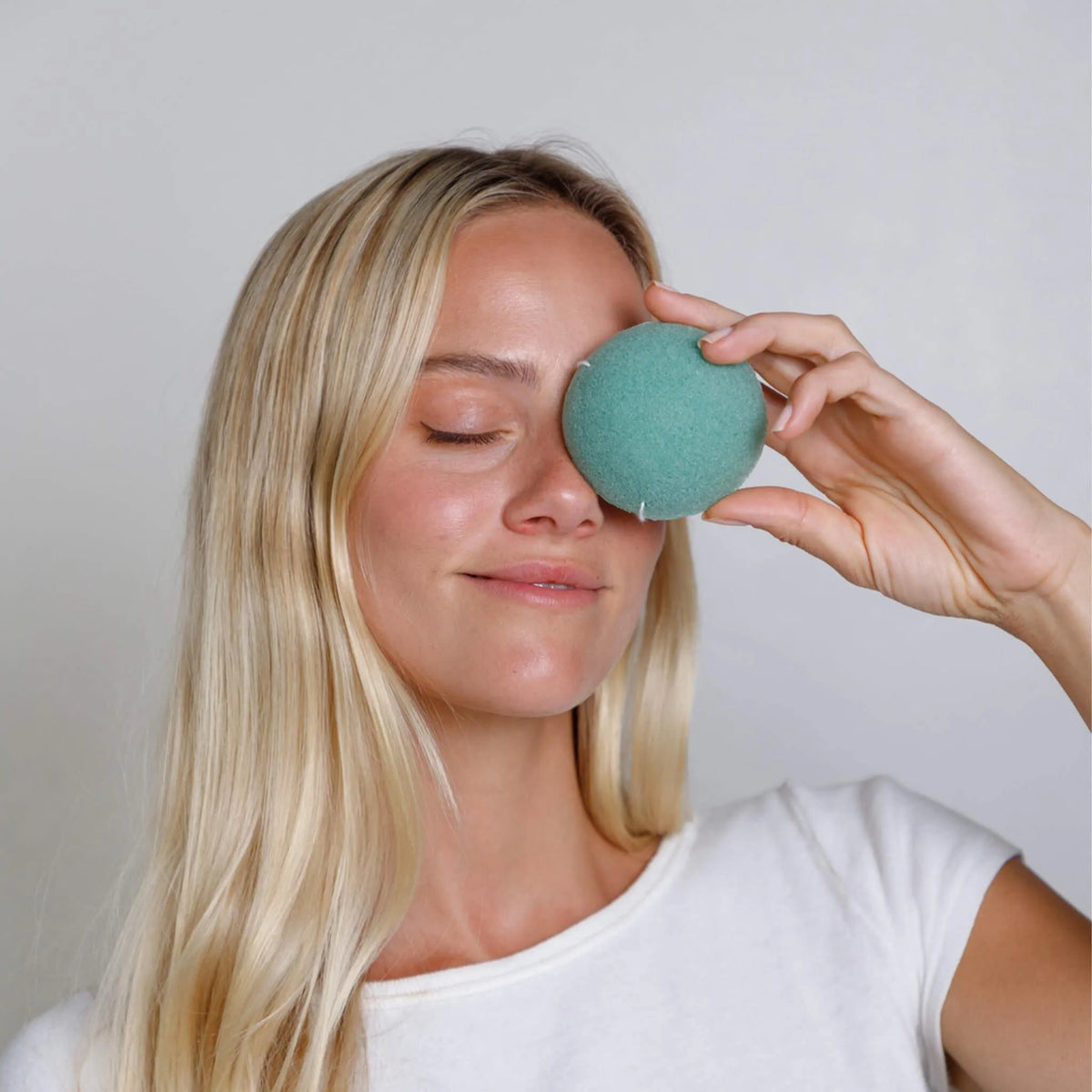 Woman holding a green face sponge to her eye against a plain background