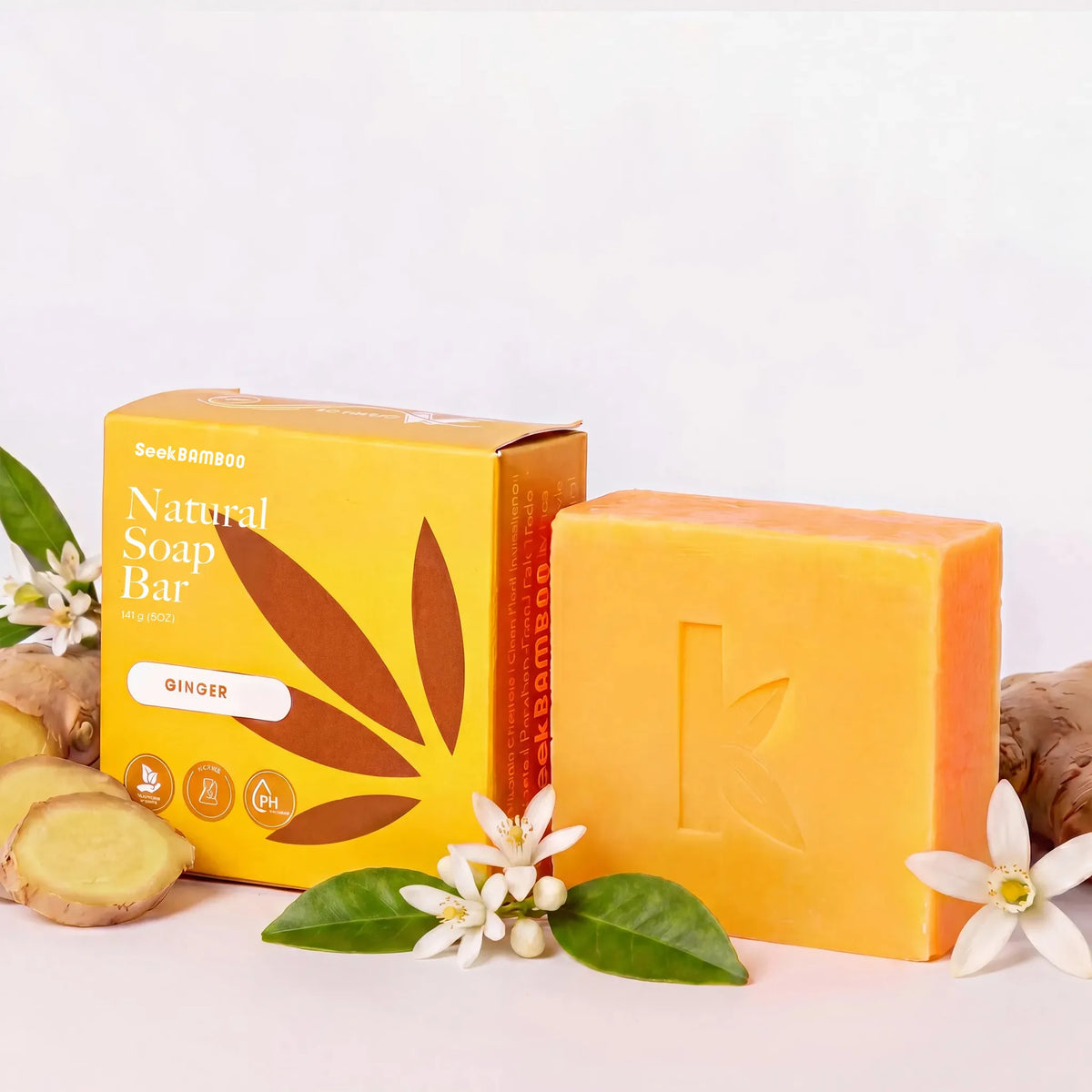 A bar of natural ginger soap and its packaging box, in warm yellow-orange tones, styled with ginger roots, green leaves, and small white flowers on a clean white background.