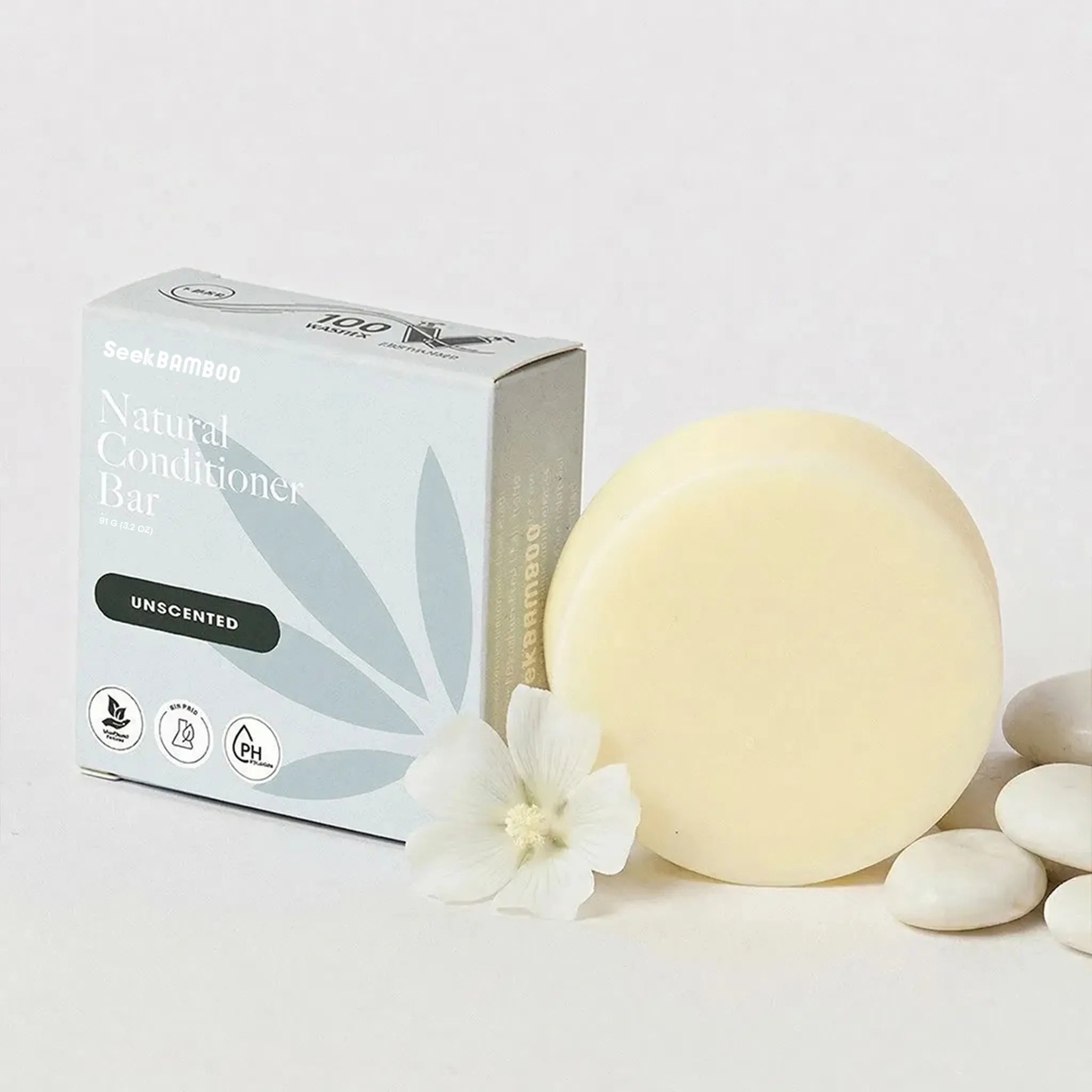 unscented conditioner bar shown with its product box, a round pale cream conditioner bar, a white flower, and smooth white stones on a clean white background.