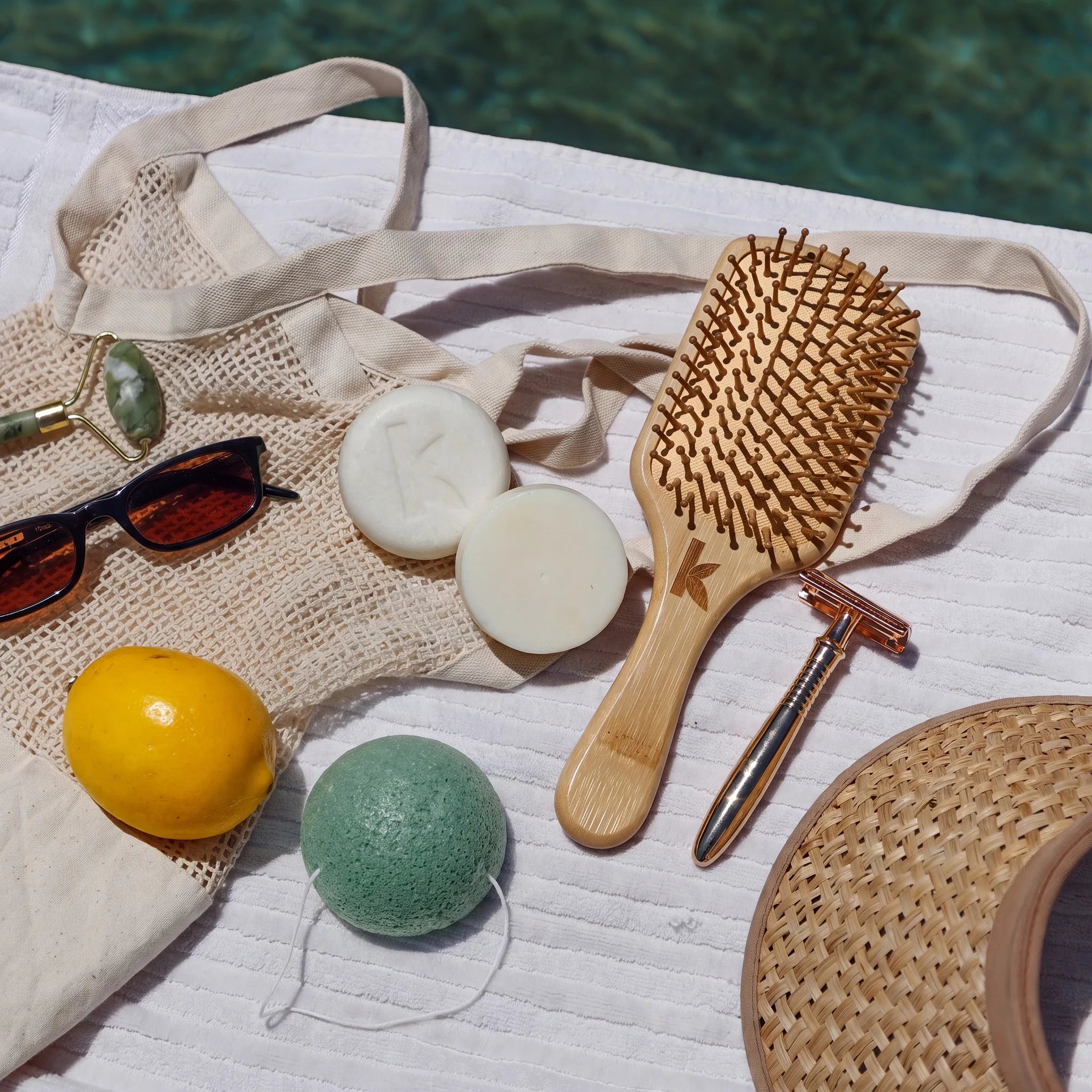 A styled flat-lay of beach and spa accessories on a white towel near a pool, including a natural wood paddle hairbrush, a woven tote bag, a yellow lemon, a green bath bomb, white soap bars, sunglasses, a rose gold safety razor, and a straw hat.