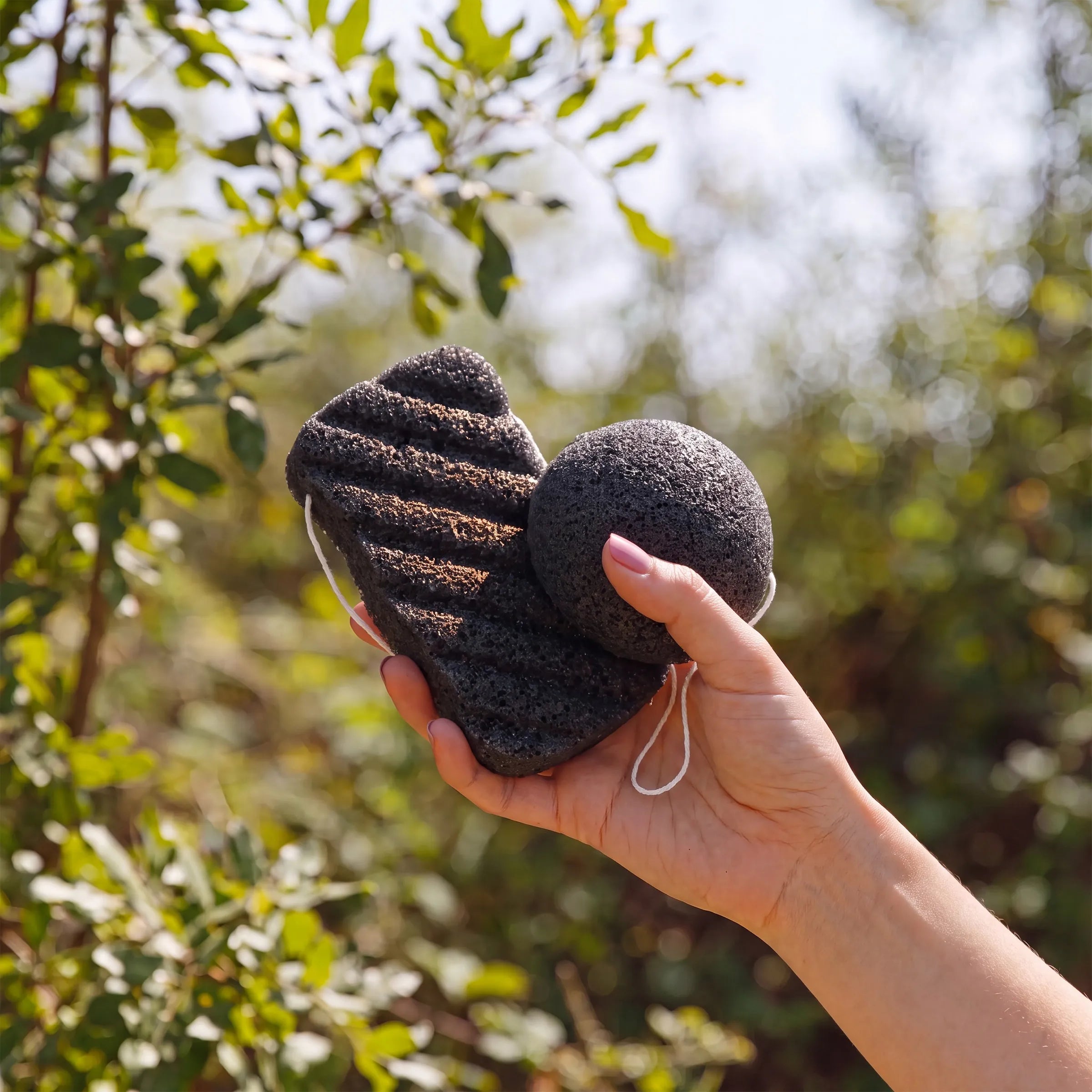 A hand holding two bamboo charcoal sponges outdoors against a blurred green foliage background.