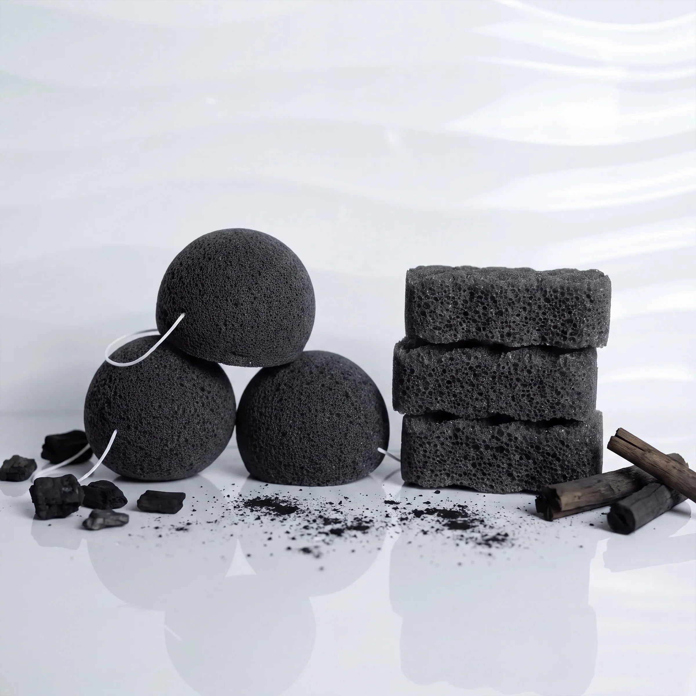 charcoal bamboo sponges with white string and three rectangular charcoal pieces stacked on a white reflective surface, with scattered charcoal powder and small wood pieces around them.