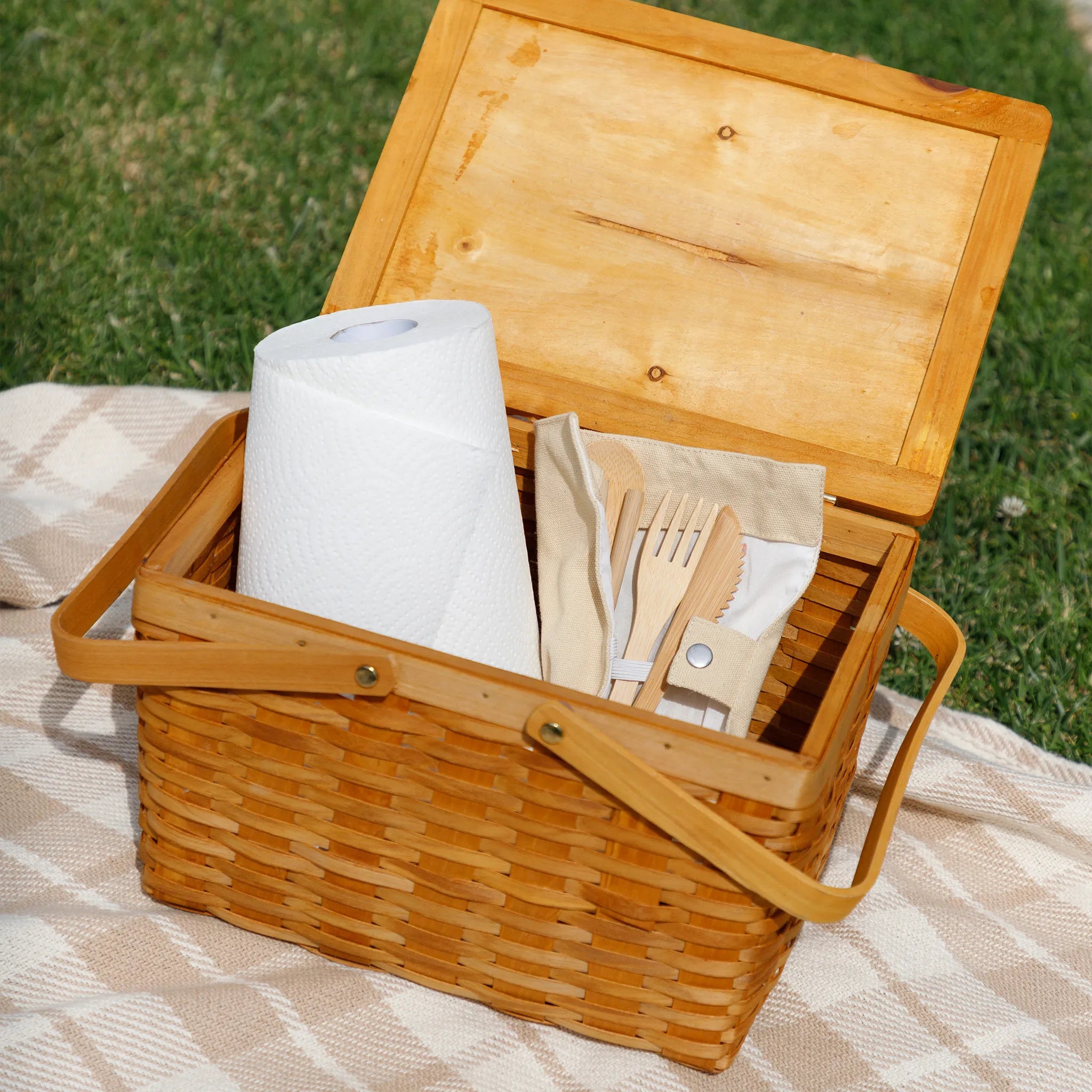 Wooden picnic basket with paper towels, bamboo cutlery, and napkins on a grassy background
