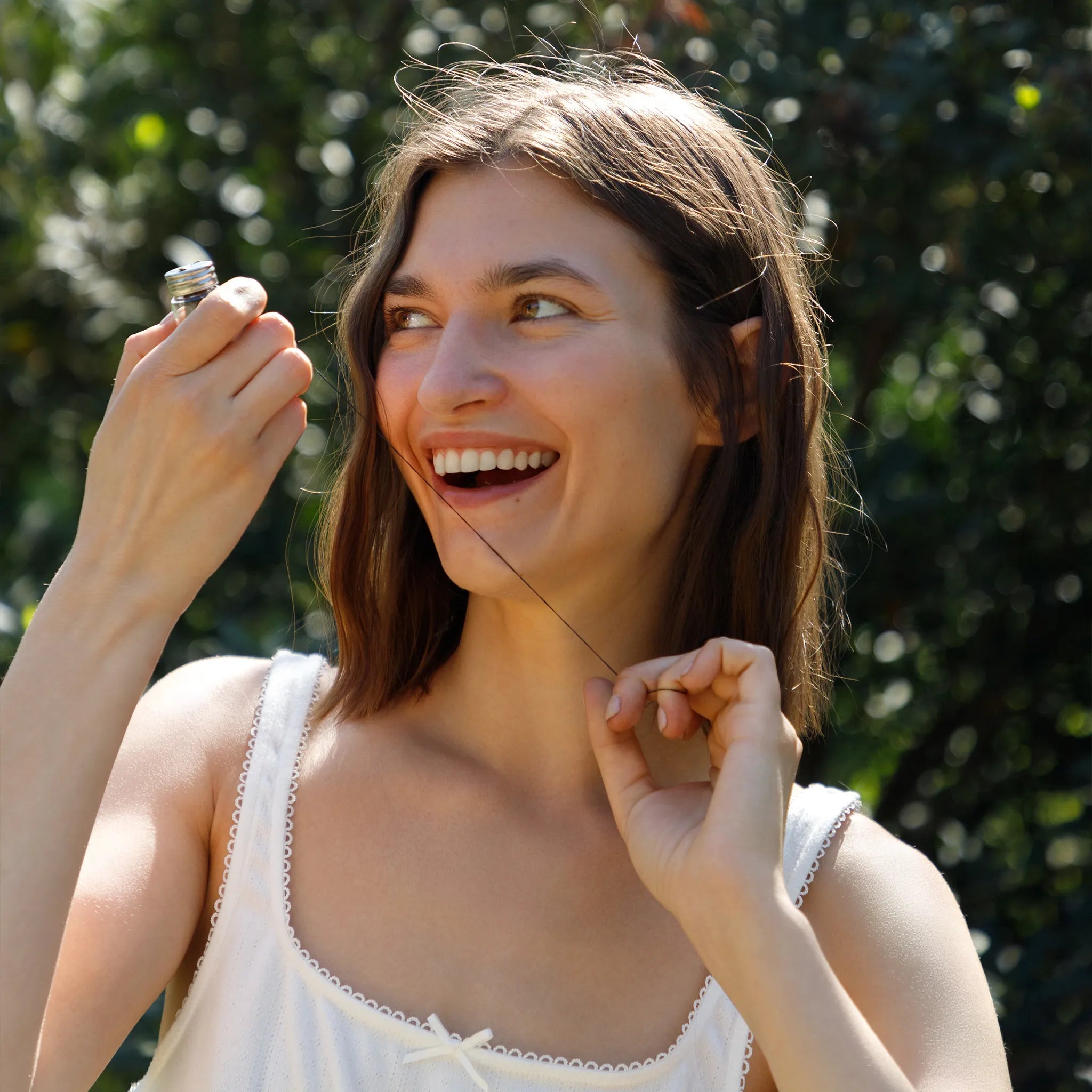 A woman smiling outdoors while holding  bamboo floss container in one hand and pulling floss with a string between her fingers with the other.