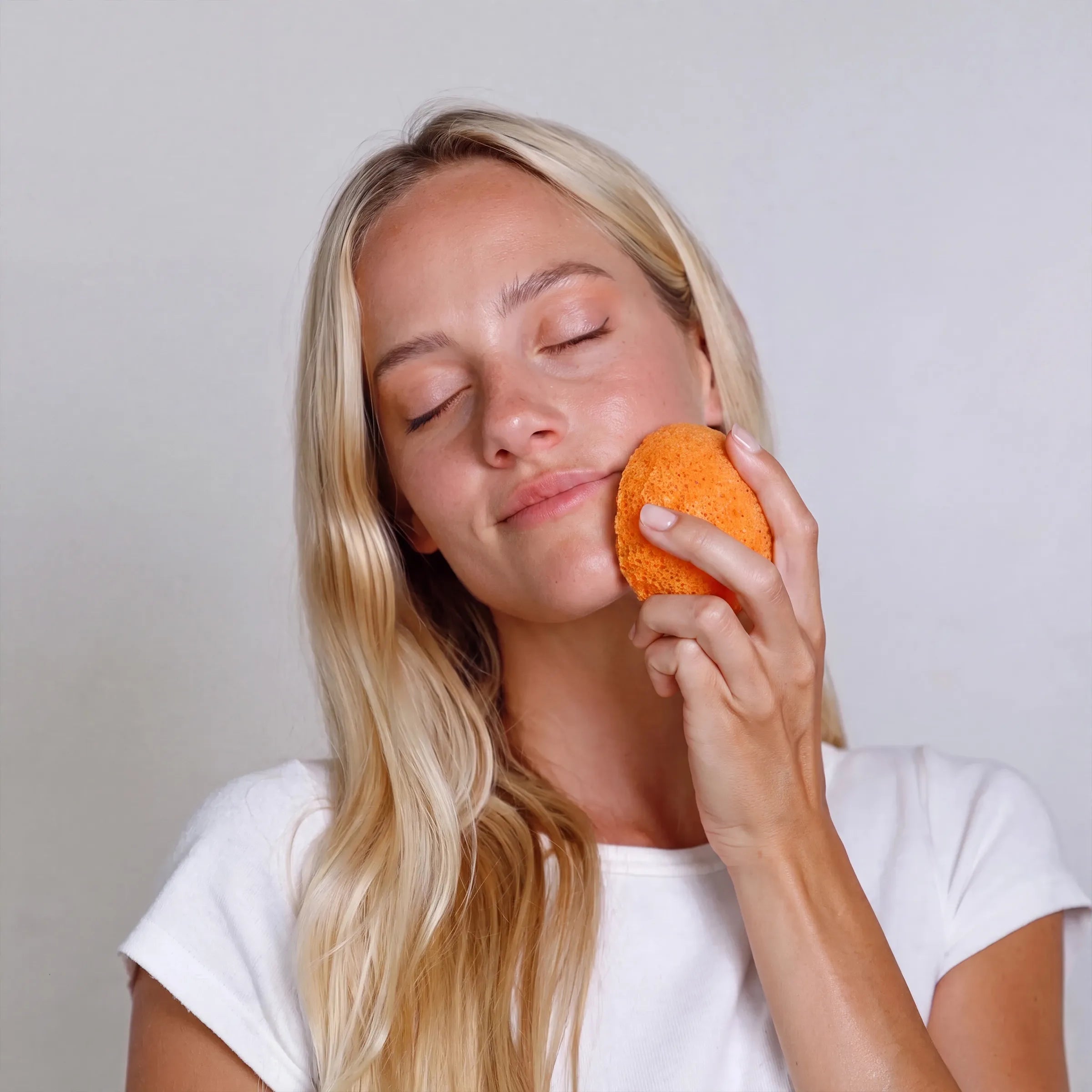 A blonde woman with her eyes closed applies an kojic acid facial sponge to her face, wearing a white t-shirt against a plain light gray background.