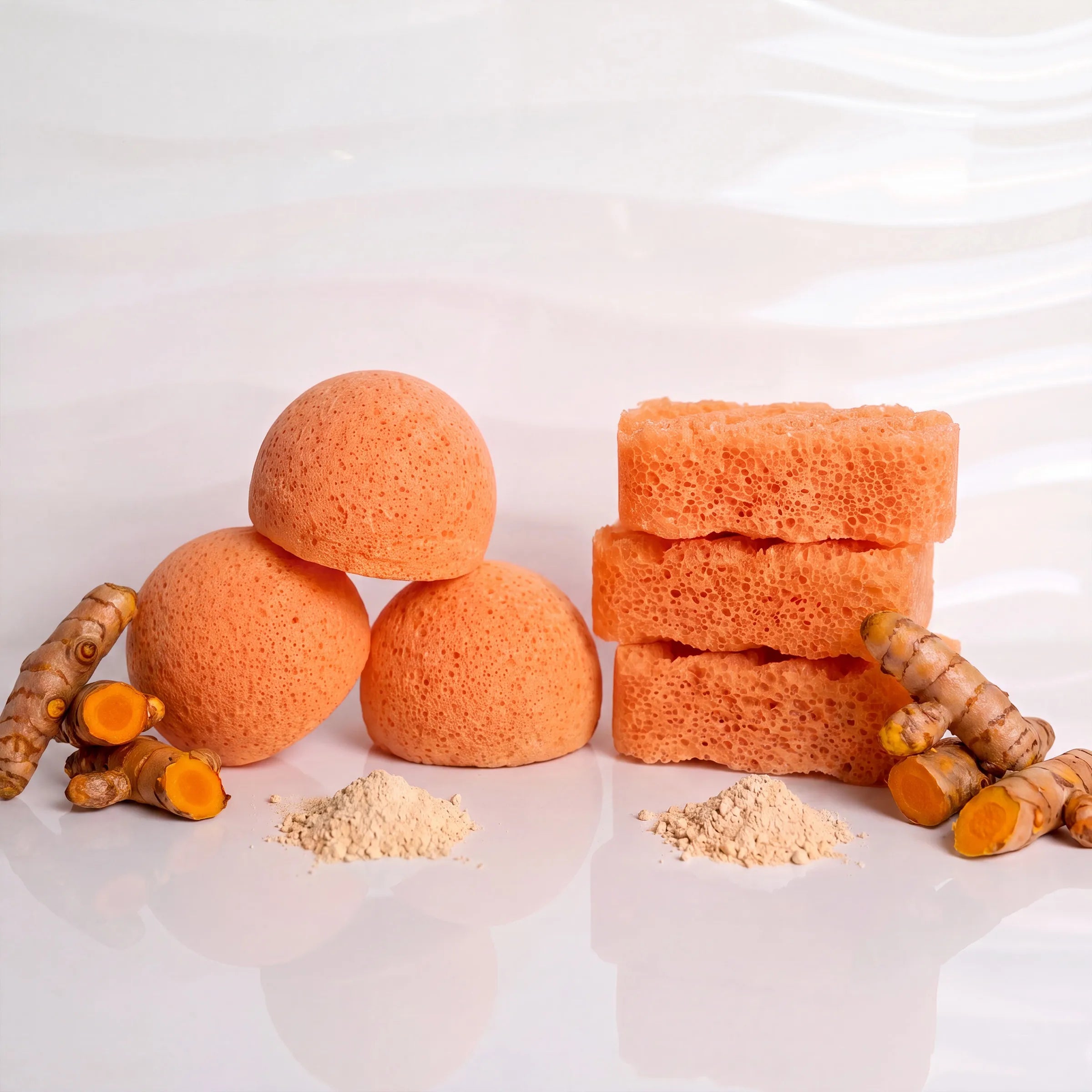 Three kogic acid konjac sponges, including two rounded bath bombs and two rectangular bath sponges, arranged with turmeric roots and small piles of powder on a glossy white reflective surface.
