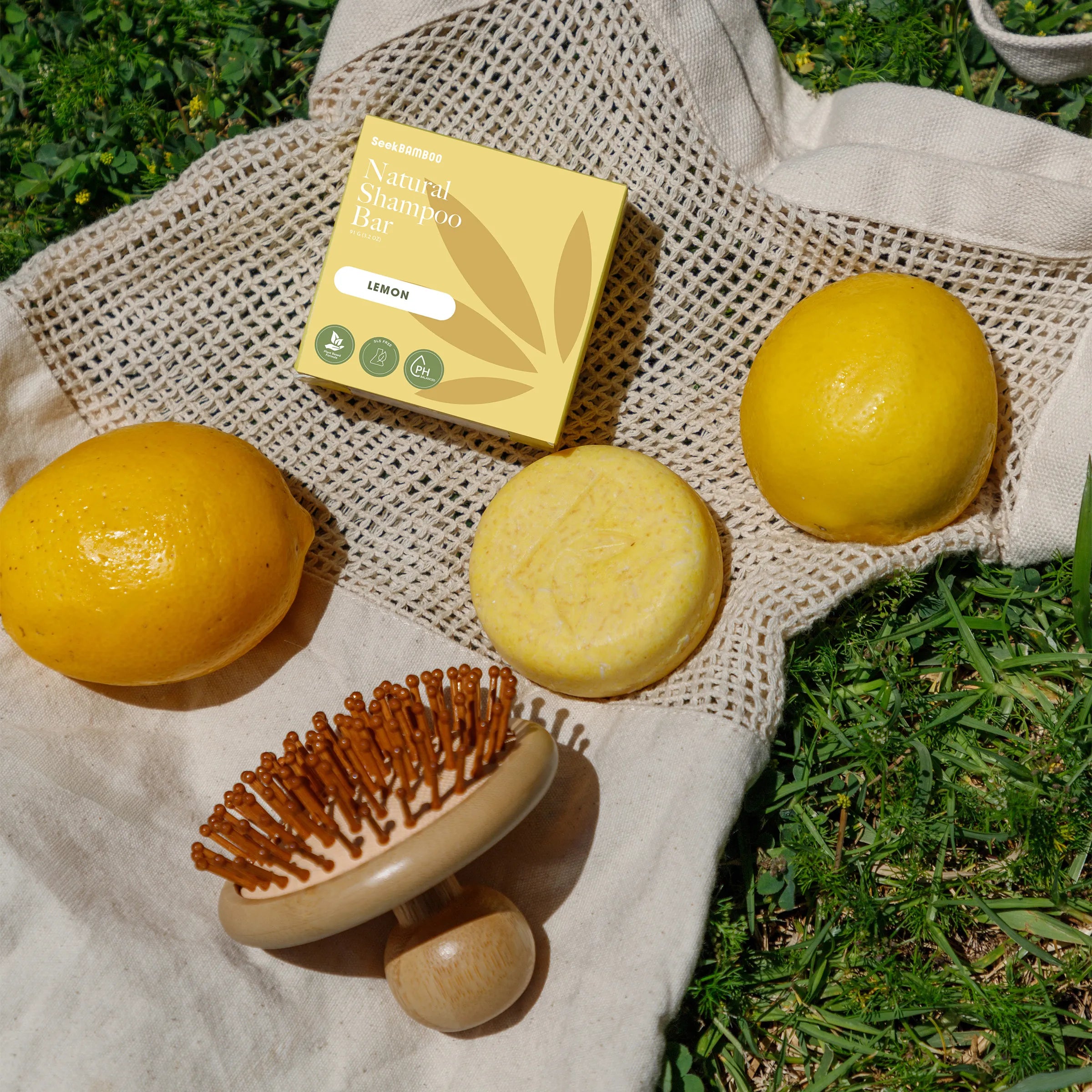 A lemon-scented shampoo bar set displayed outdoors on a beige mesh bag over green grass. The set includes a yellow box labeled 'sanitary Shampoo Bar' with 'LEMON' on it, a round yellow shampoo bar, a yellow lemon, a wooden-handled brush with brown bristles, and a small yellow pouch.