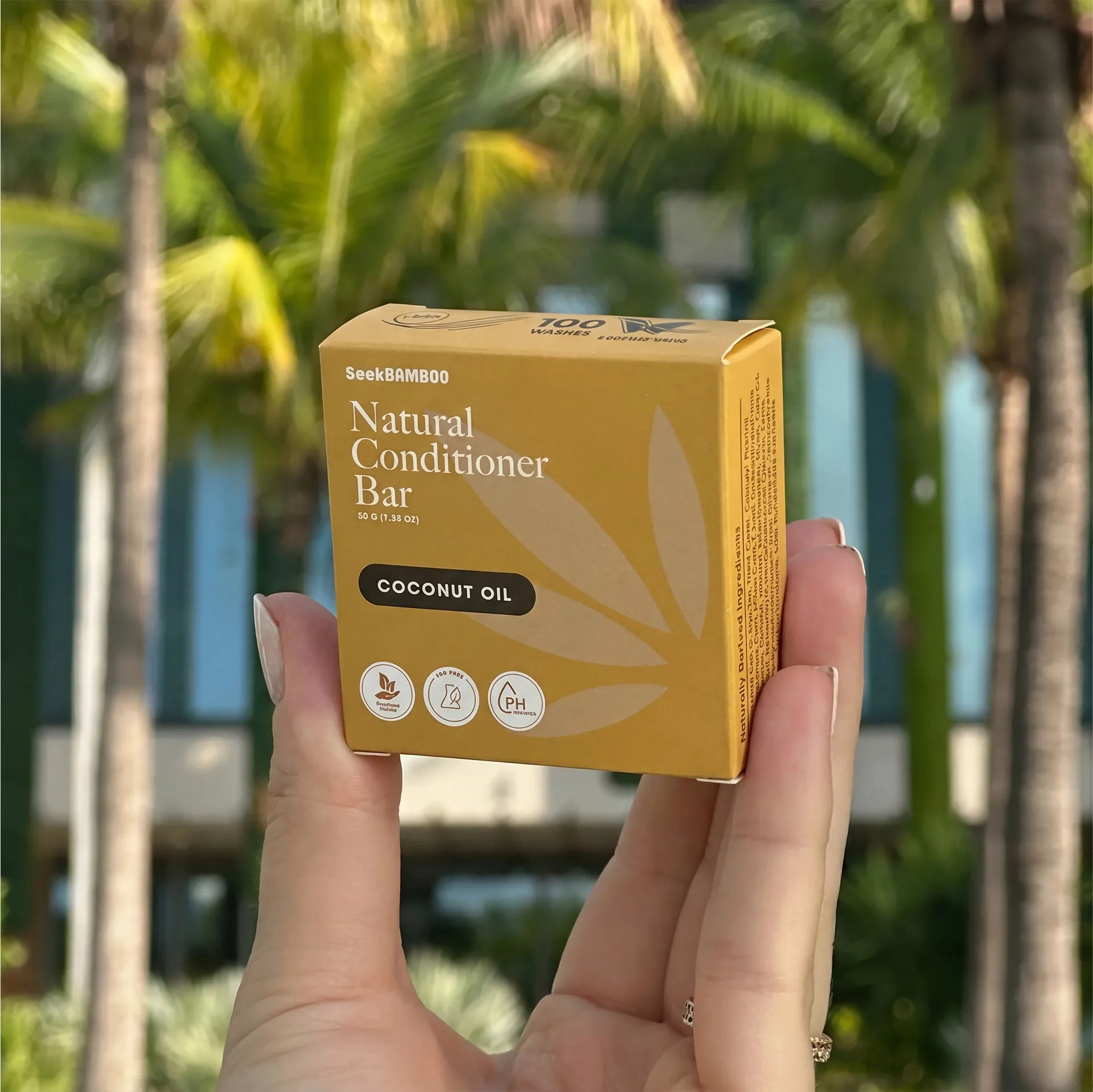 Hand holding a box of Seek Bamboo Natural Conditioner Bar with coconut oil in front of palm trees.