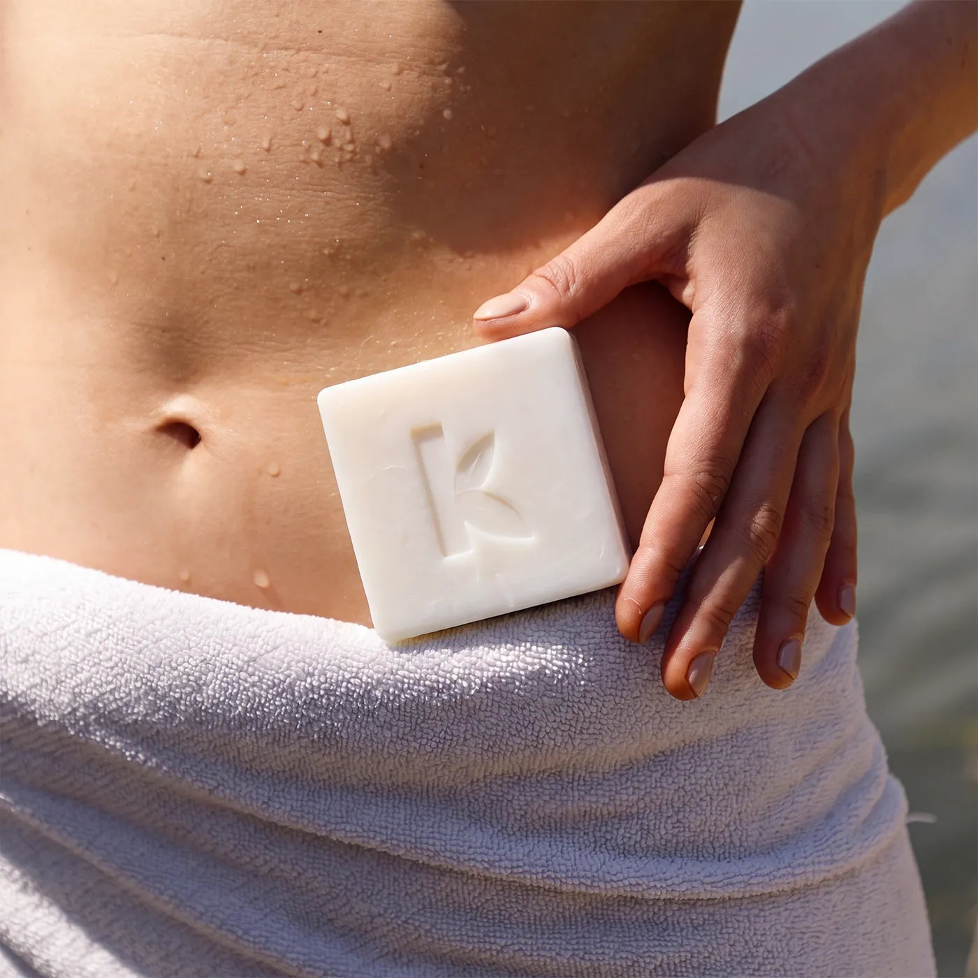 A close-up lifestyle shot of a person wrapping a white towel around their waist while holding a coconut oil bar of soap with an embossed leaf logo against their lower abdomen.