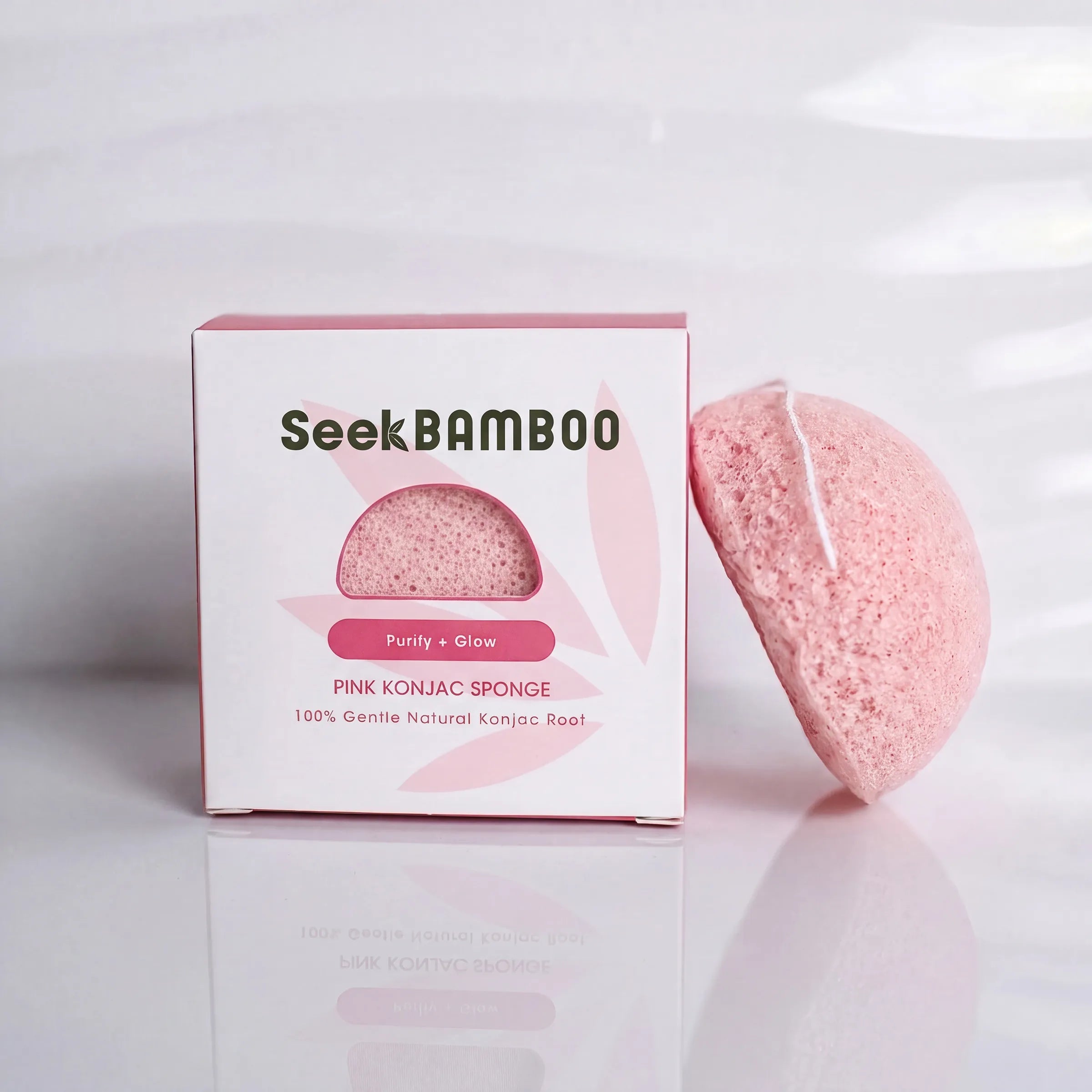A pink konjac sponge makeup remover by SeekBAMBOO, shown next to its white and pink retail box on a reflective white surface.