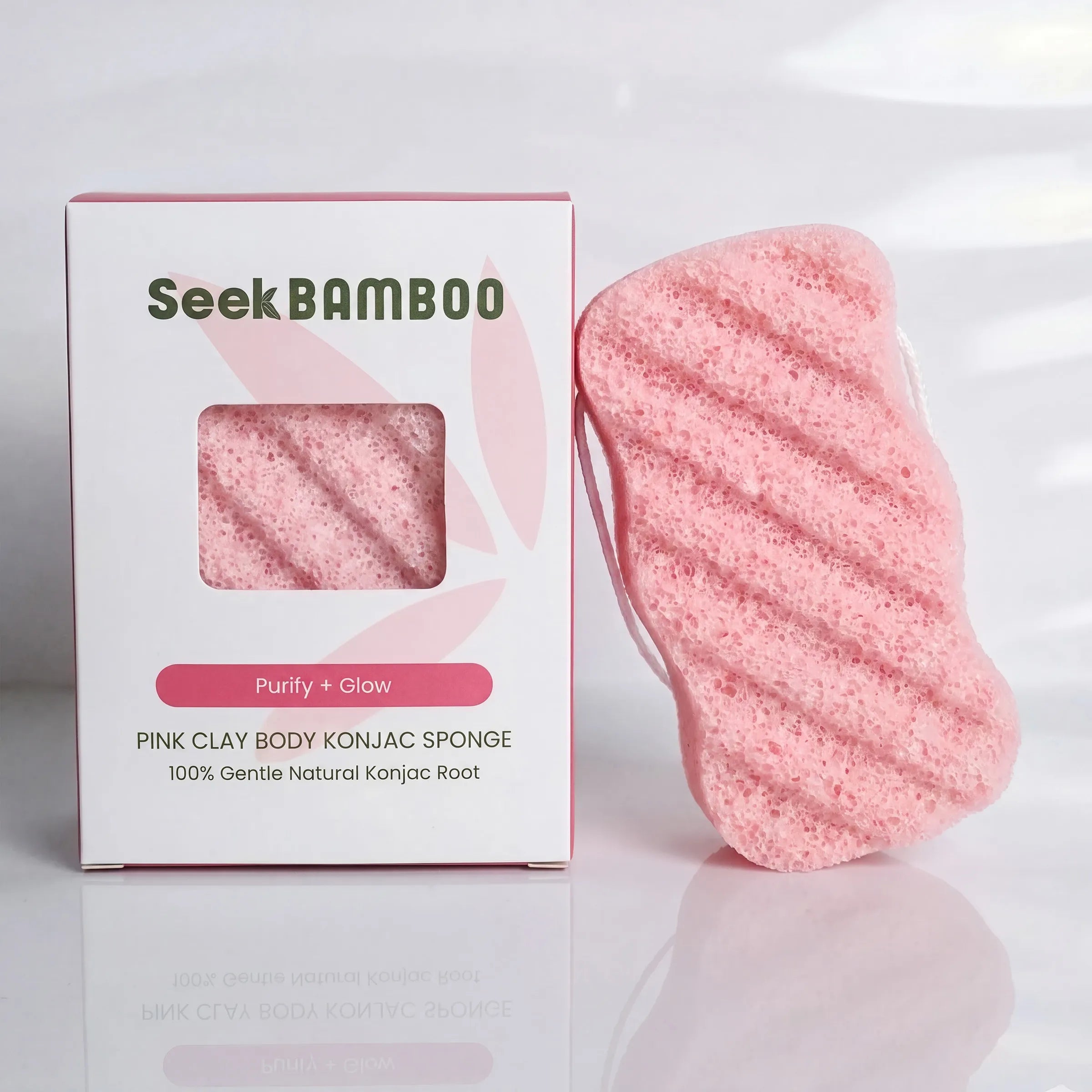 A pink clay body konjac sponge next to its white and pink packaging box, branded SeekBAMBOO, on a clean white reflective surface.