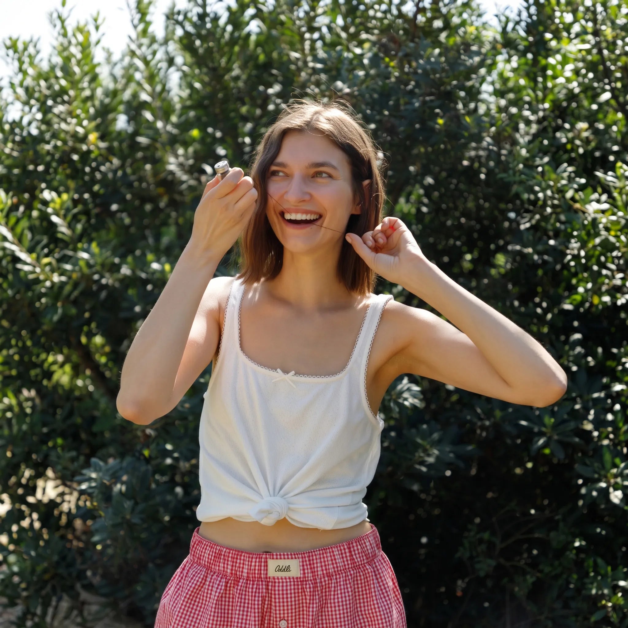 A woman outdoors in front of green foliage, wearing a white tank top tied at the waist and red-and-white gingham shorts. She is smiling and posing with bamboo floss.