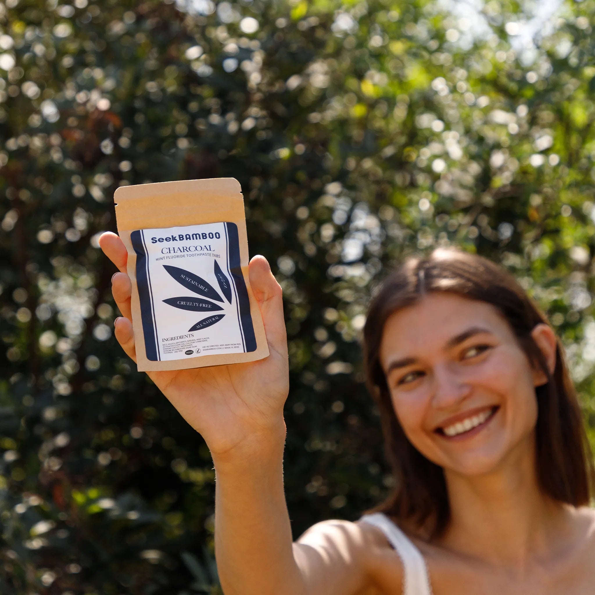 A woman outdoors holds up a pouch of Soak Bamboo Charcoal toothpaste tablets, with a blurred green garden background.