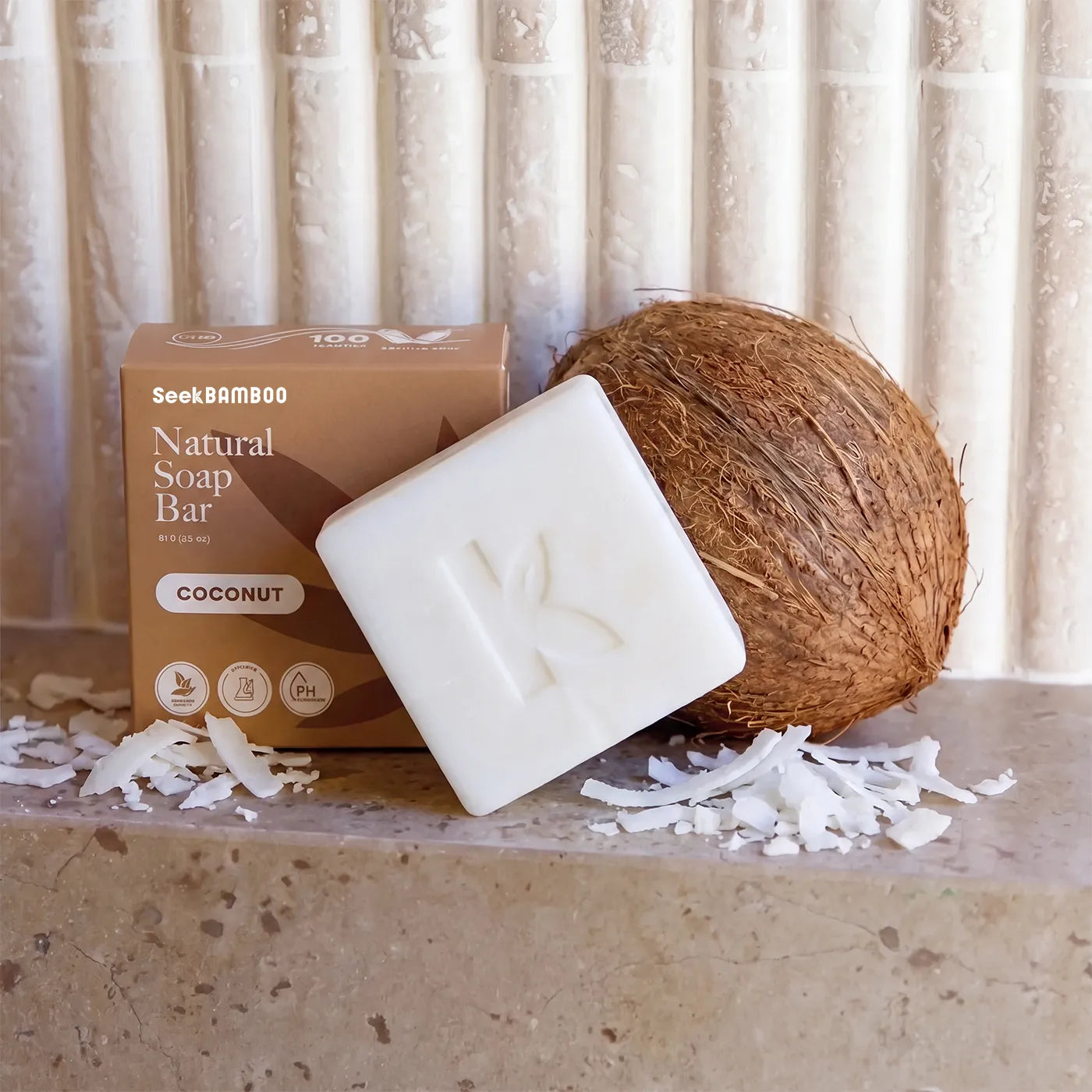 A bar of coconut soap from SeekBAMBOO shown with its brown cardboard box, a whole coconut, and scattered coconut flakes on a stone surface against a tiled wall.