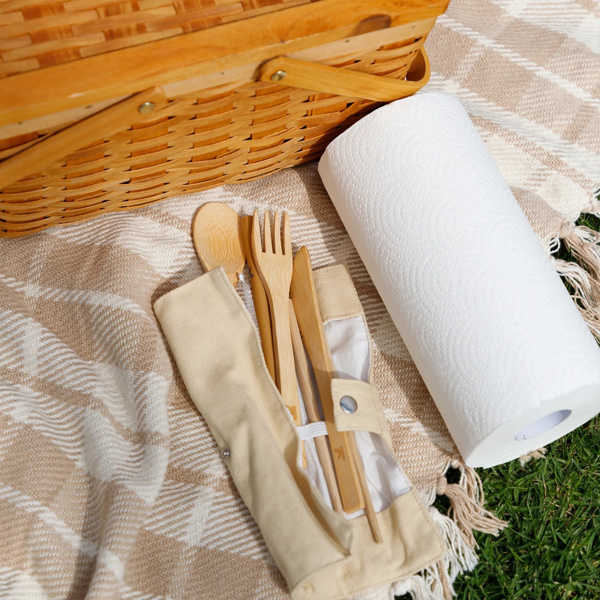Picnic setup with wicker basket, paper towels, and bamboo cutlery on a blanket outdoors.