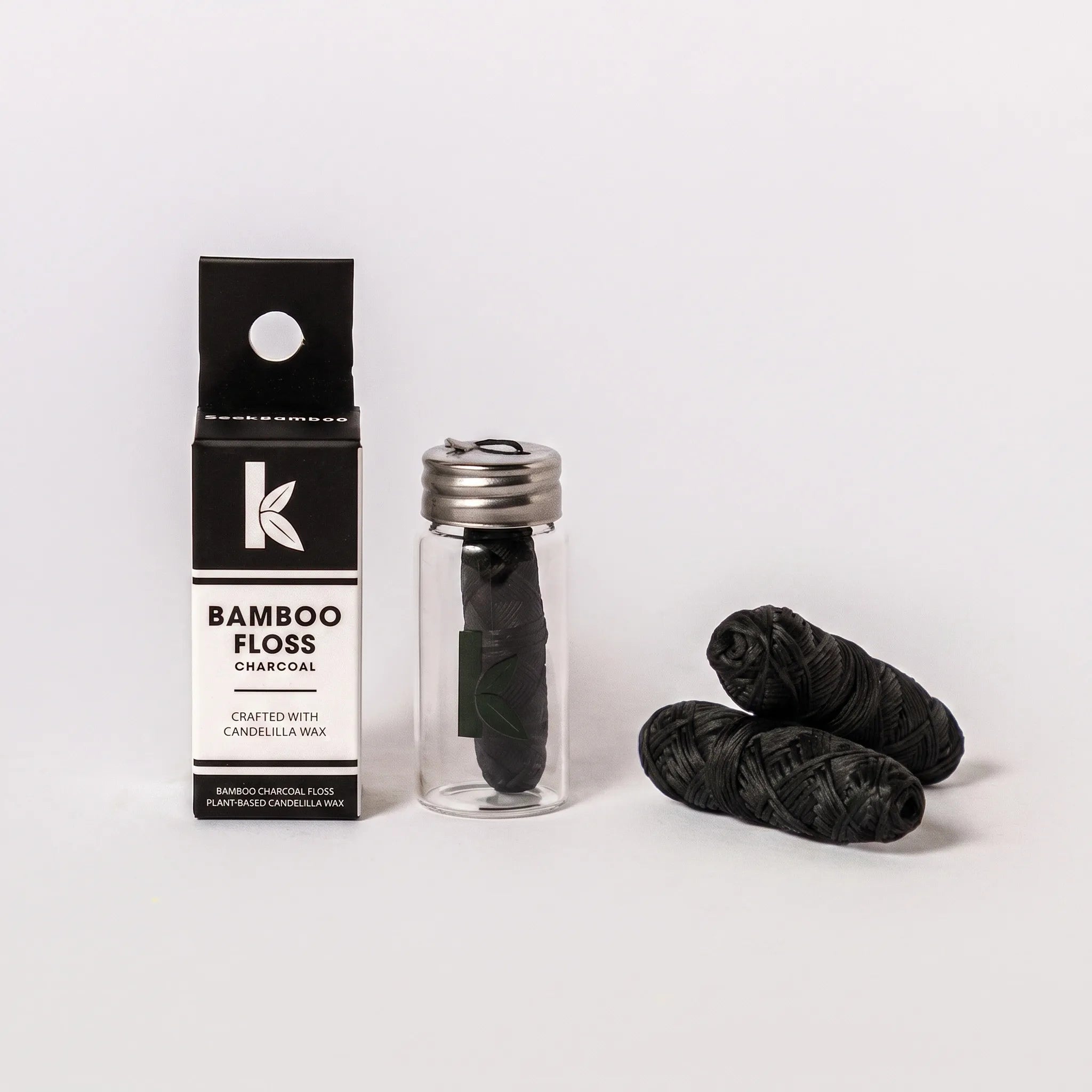 Bamboo floss charcoal packaging shown with a seek bamboo box, a clear glass bottle with a silver cap, and three dark charcoal floss balls on a light gray background.